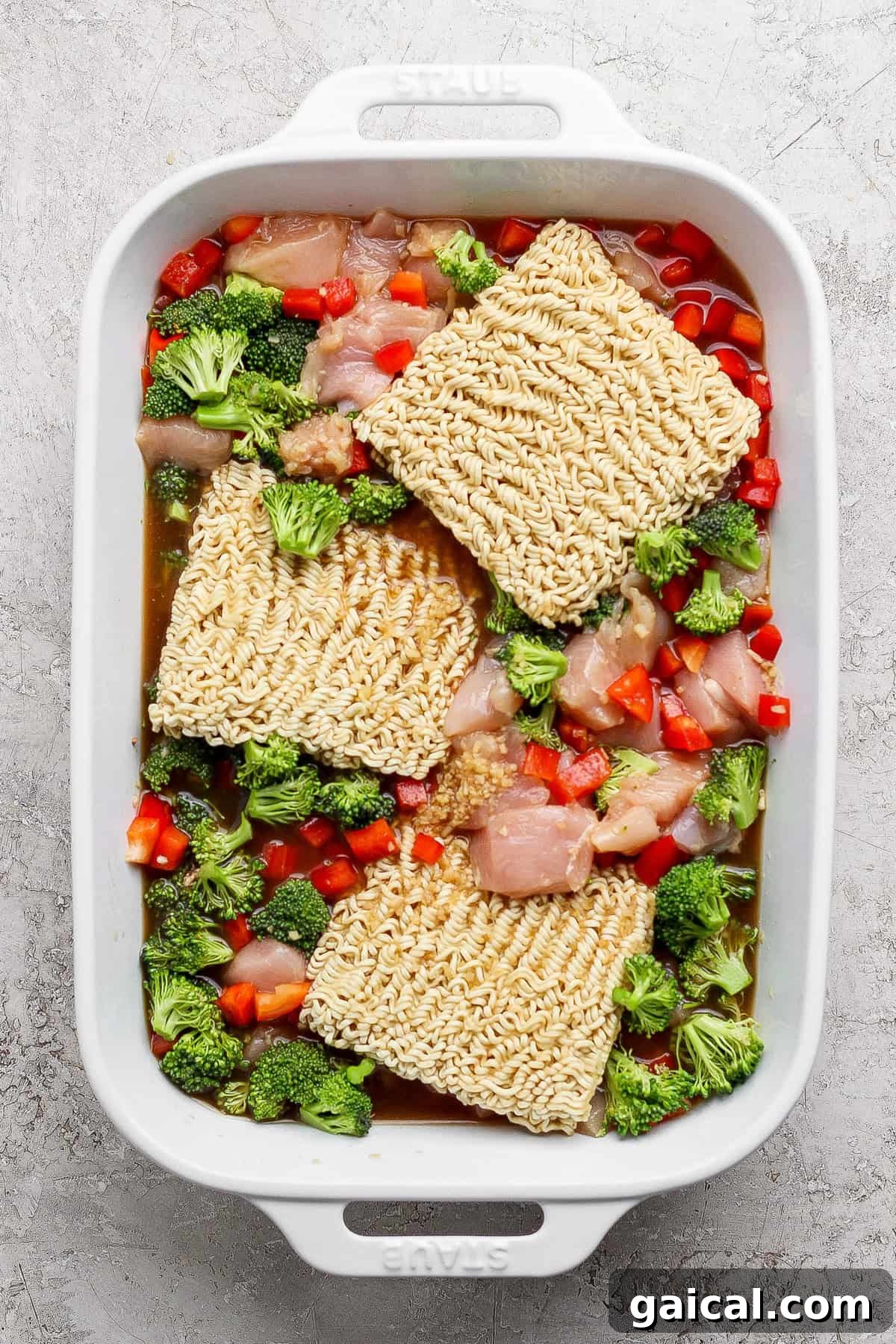Uncooked ramen noodles, raw chicken pieces, chopped broccoli, and diced red bell peppers in a white rectangular baking dish.