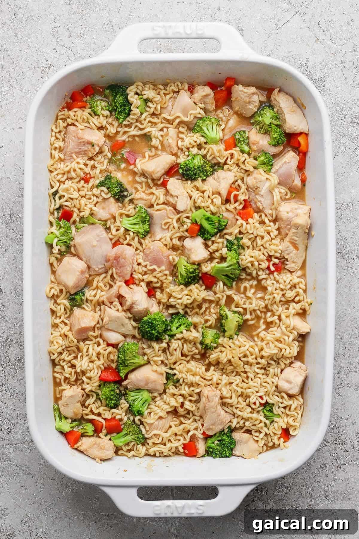 A white baking dish filled with cooked ramen noodles, pieces of chicken, broccoli florets, and red bell pepper in a light broth.