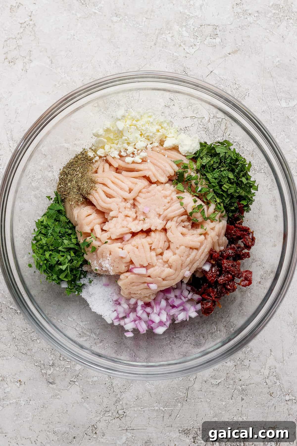 A glass bowl containing ground meat, chopped red onion, herbs, minced garlic, sun-dried tomatoes, salt, pepper, and seasonings arranged separately.