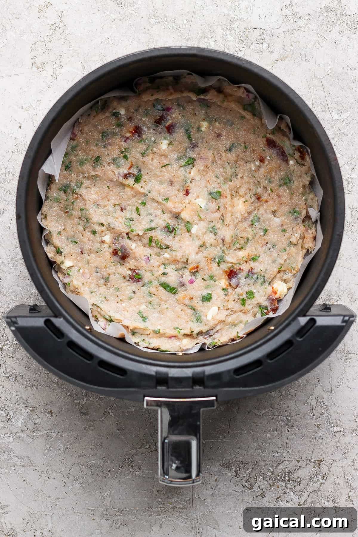 Uncooked mixture of herbs, vegetables, and cheese pressed into a round baking dish lined with parchment paper inside an air fryer basket.