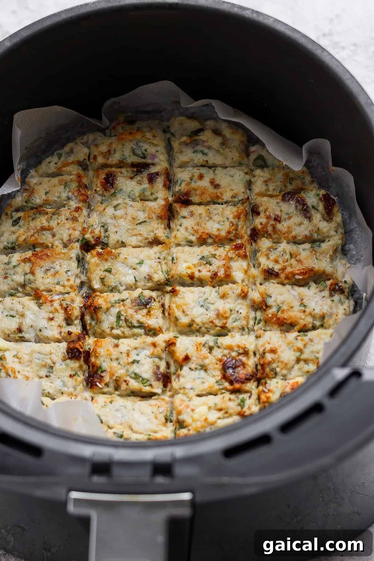 A round dish of baked, sliced savory bars with herbs and browned spots, lined with parchment paper, inside an air fryer basket.
