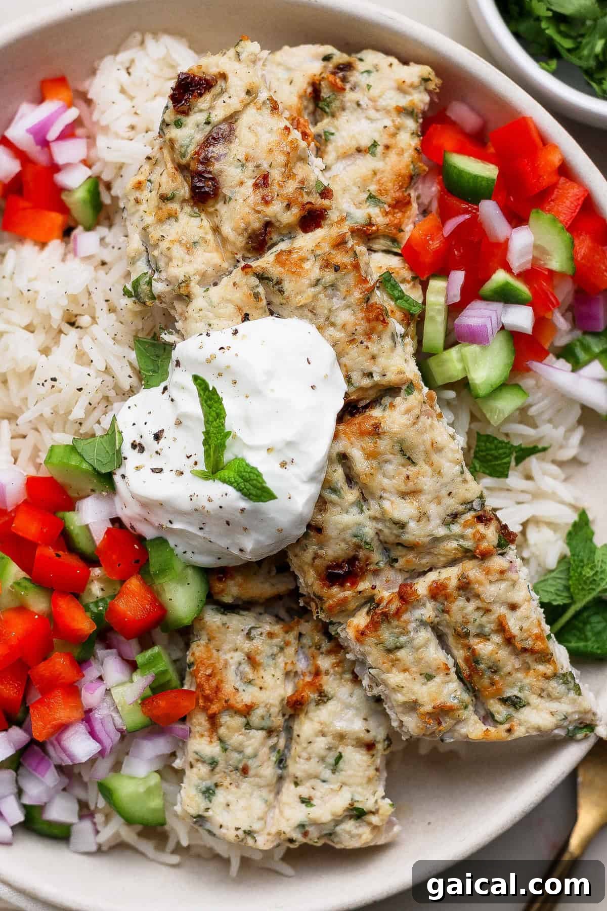 A plate of rice topped with grilled chicken kebabs, chopped red onion, red bell pepper, cucumber, and a dollop of yogurt, garnished with a mint leaf.