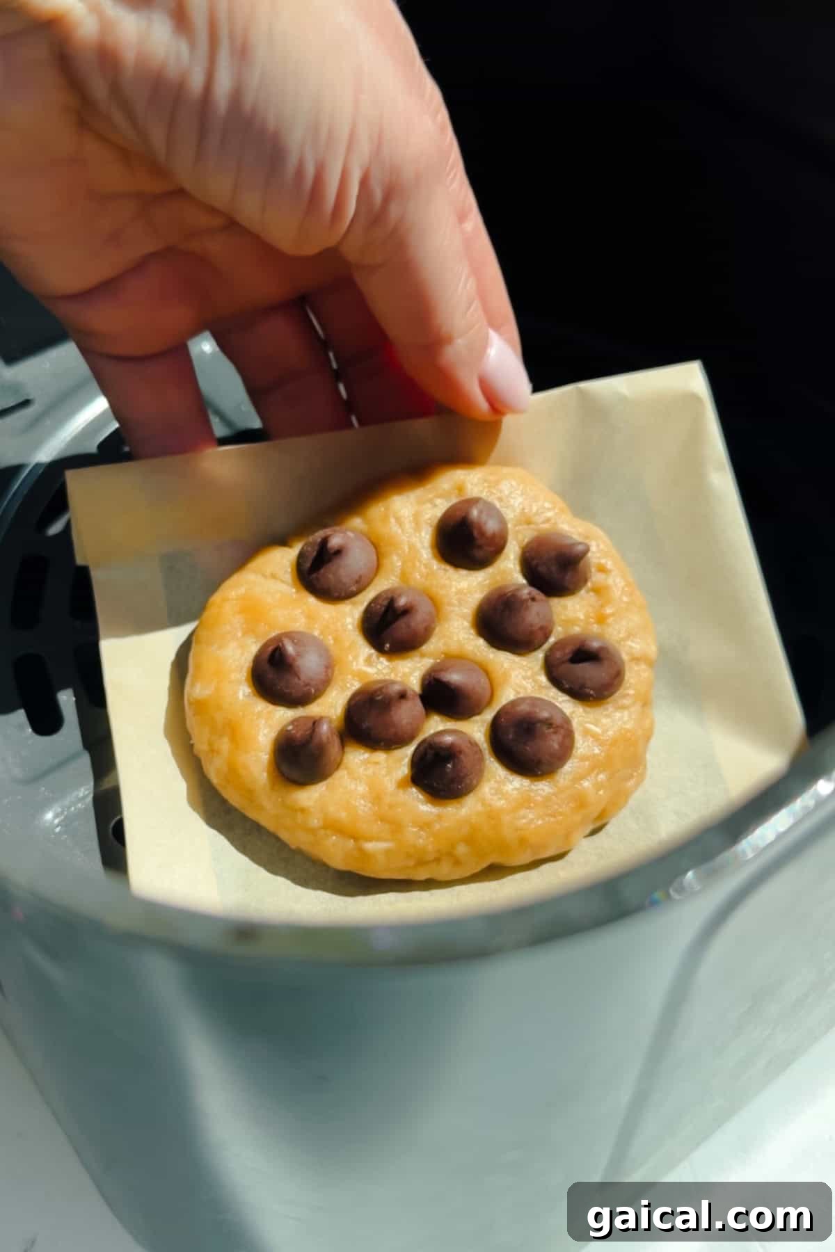 A hand is placing a round chocolate chip cookie dough on a piece of parchment paper inside an air fryer basket.