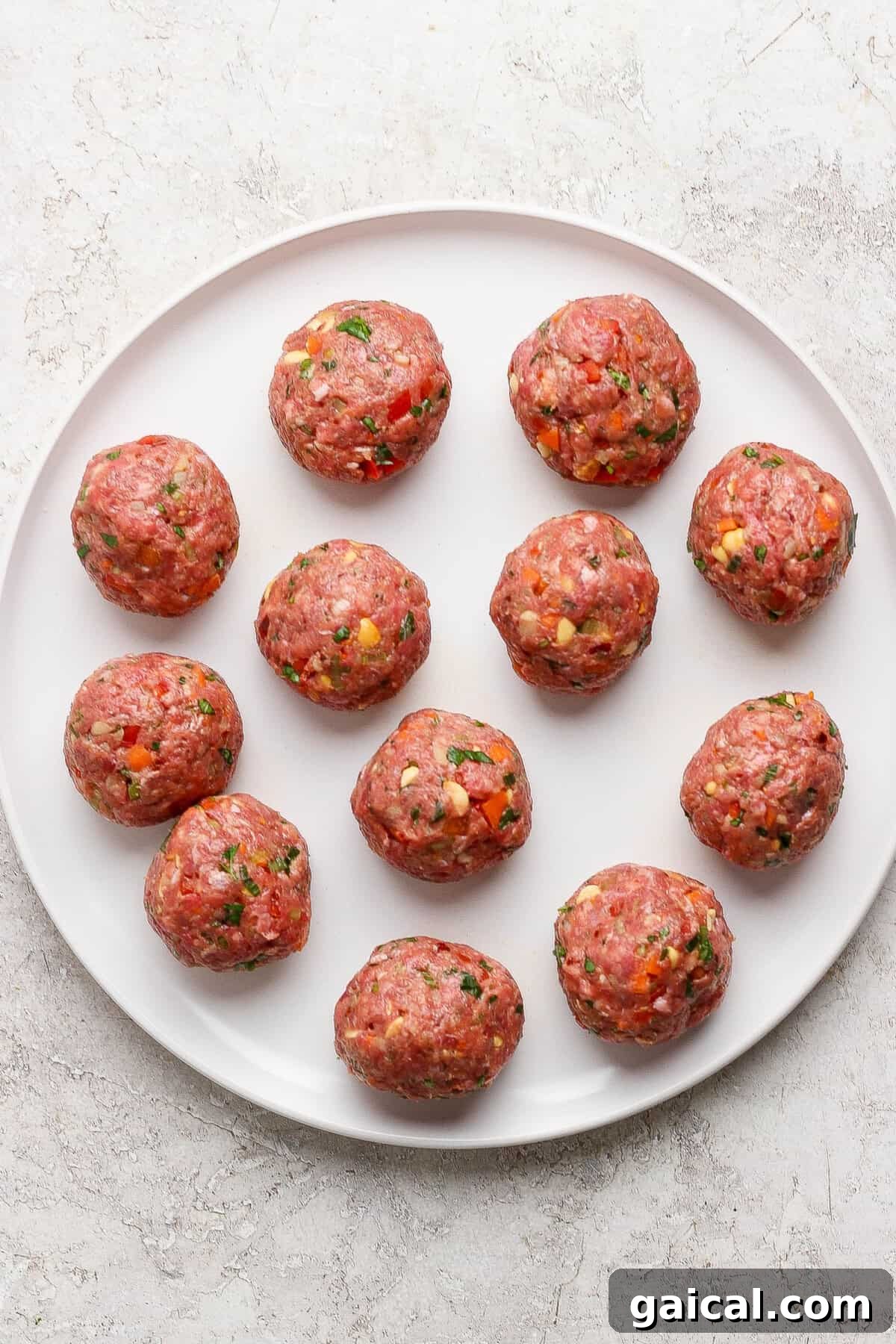 A white plate with twelve uncooked meatballs, mixed with herbs and small vegetable pieces, on a light textured surface.