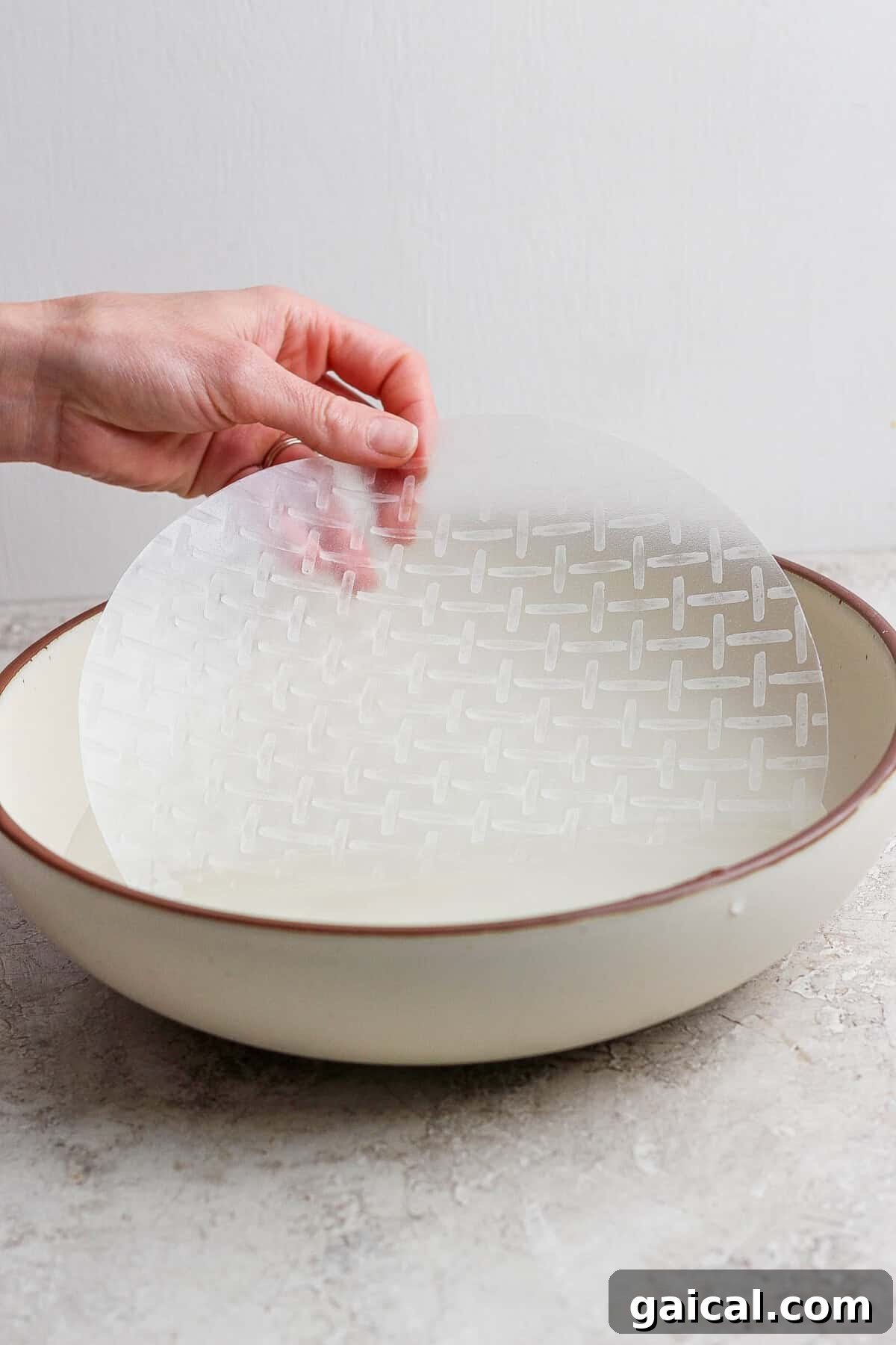 A hand dips a round sheet of rice paper into a shallow bowl of water on a light-colored surface.