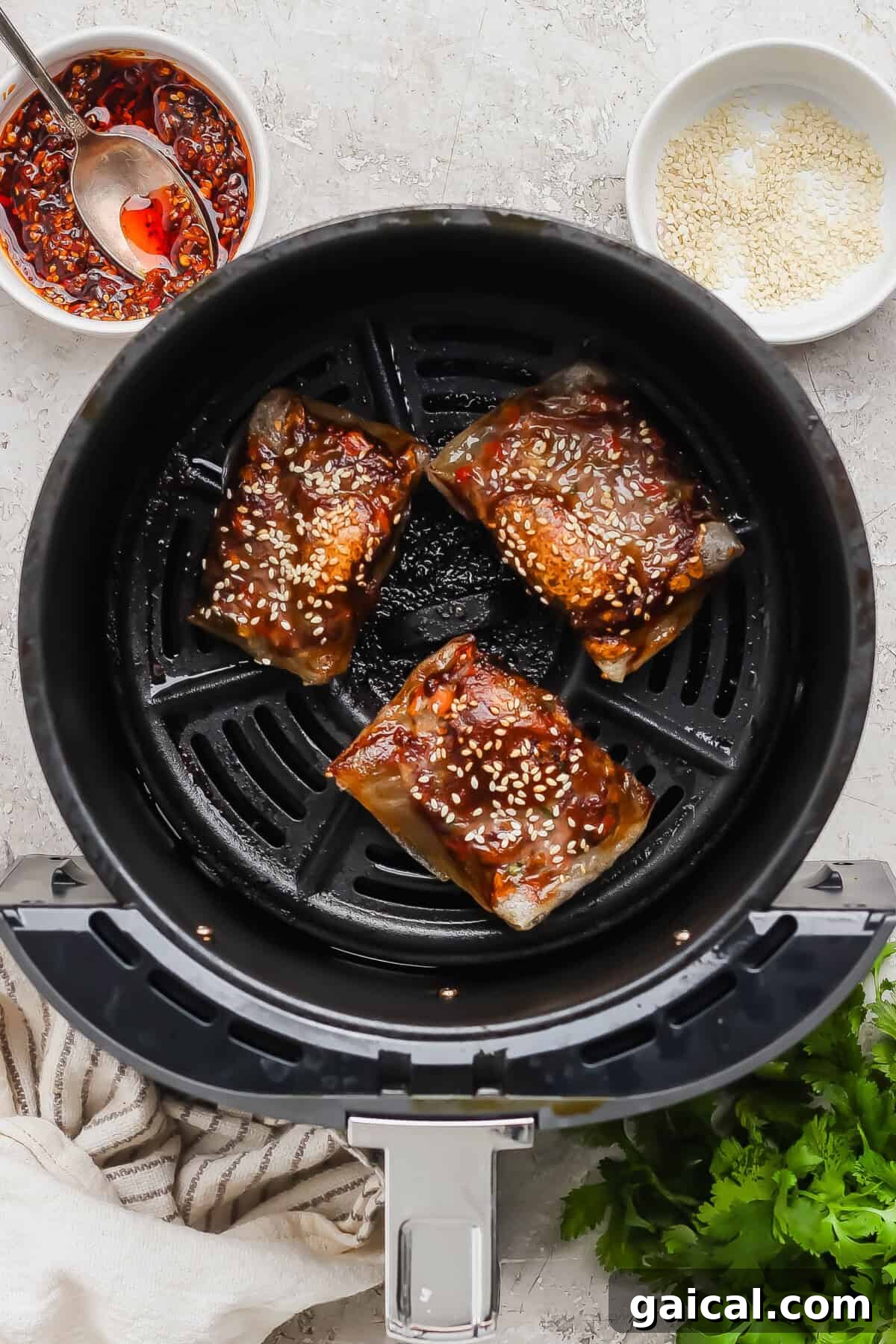Three pieces of marinated fish with sesame seeds in an air fryer basket, with a bowl of chili sauce and a bowl of sesame seeds nearby.