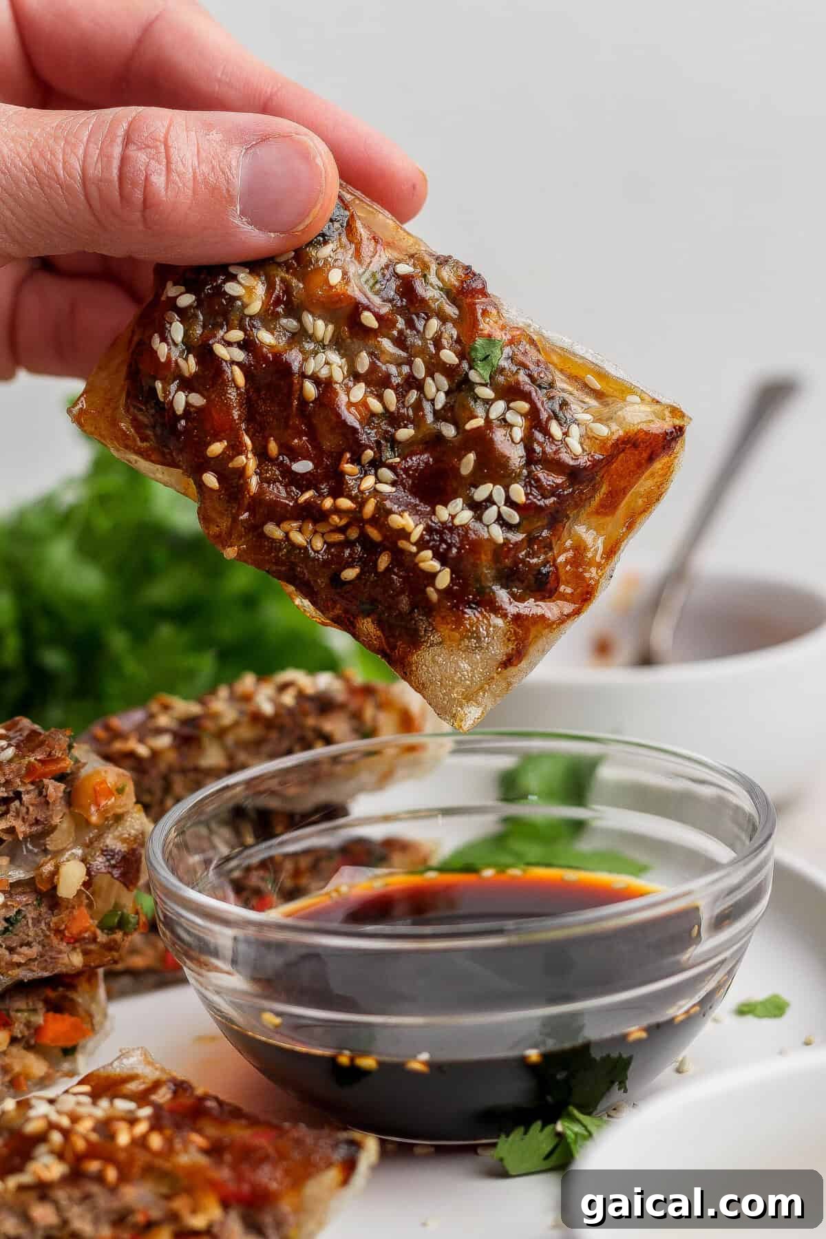A hand holds a crispy, rectangular pastry topped with sesame seeds, about to dip it into a bowl of soy sauce; greens and more pastries are in the background.