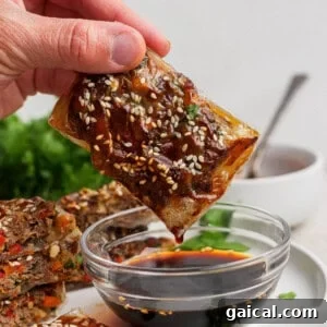 A hand dips a rectangular, sesame seed-topped pastry into a bowl of dark soy sauce. Other similar pastries and green herbs are visible in the background.