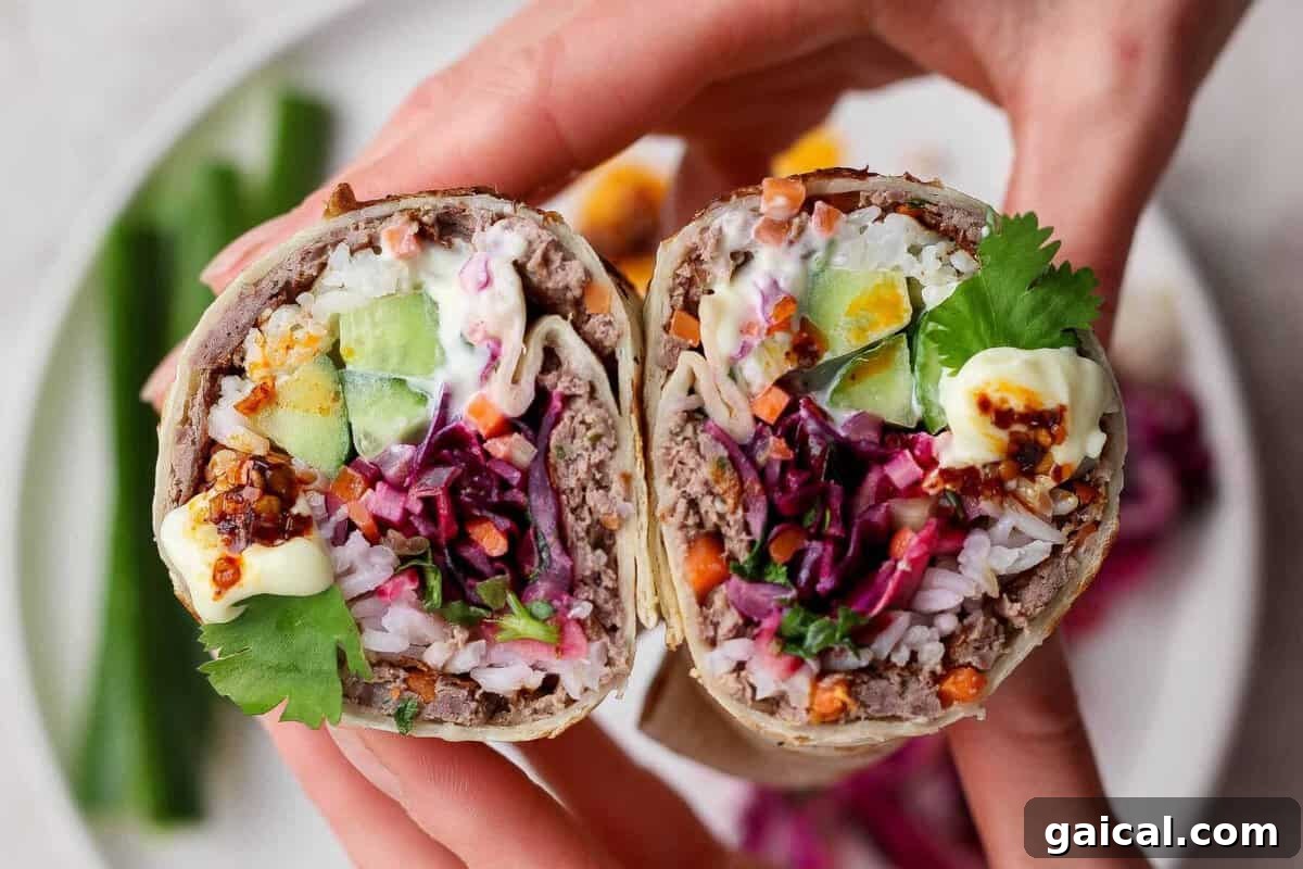 A close-up of hands holding a cut burrito filled with rice, ground meat, cucumber, egg, shredded vegetables, cilantro, and sauce, with a plate of food in the background.