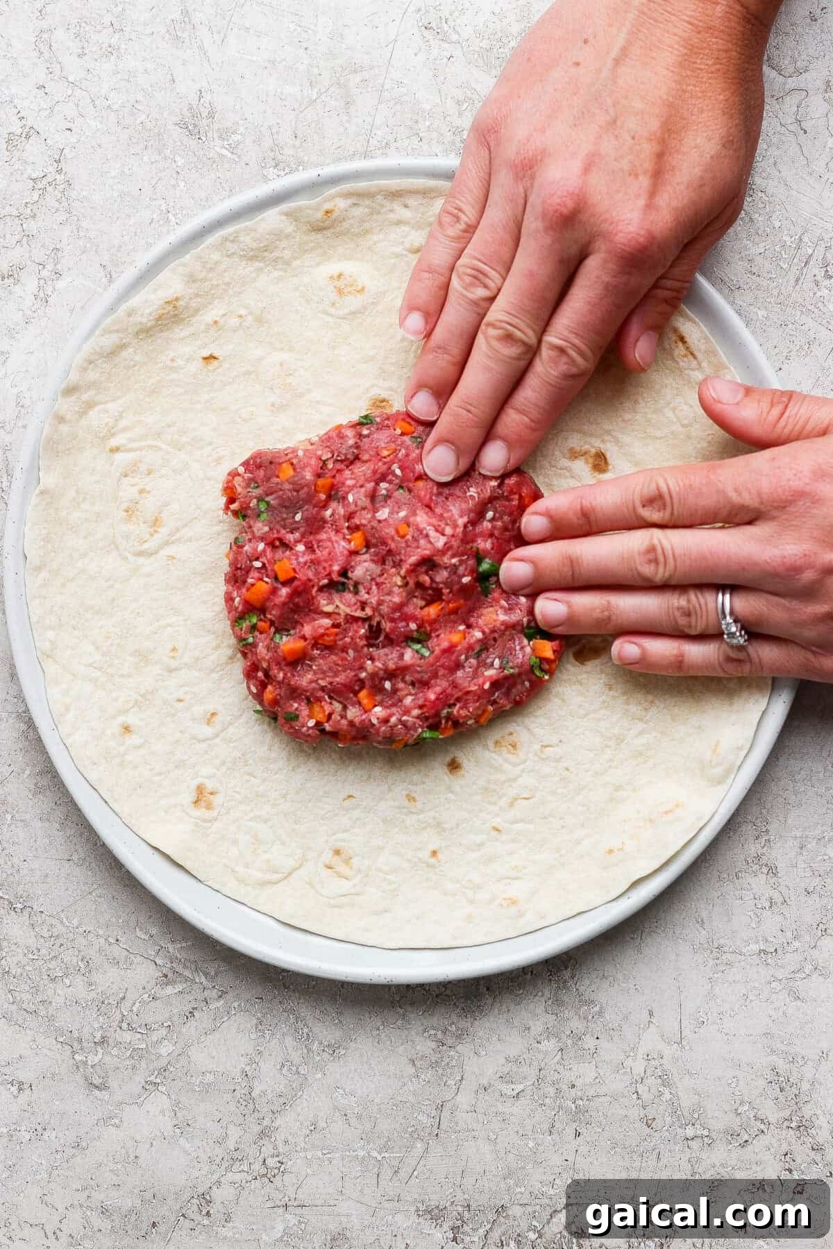 Hands pressing raw ground meat mixture with chopped vegetables onto a large tortilla on a plate.