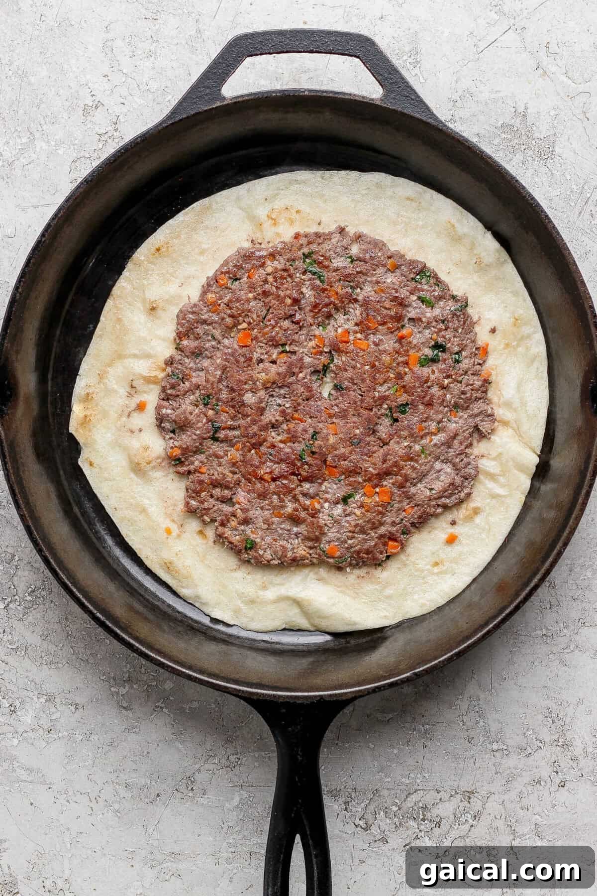 A cast iron skillet with flatbread topped with a spread of seasoned ground meat and chopped vegetables.