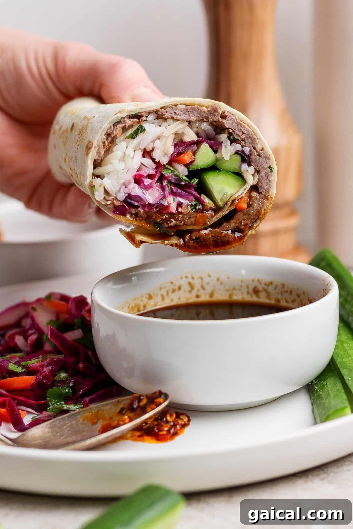 A hand holds a partially eaten wrap filled with rice, vegetables, and sauce above a white bowl of dipping sauce on a plate with salad and cucumber slices.