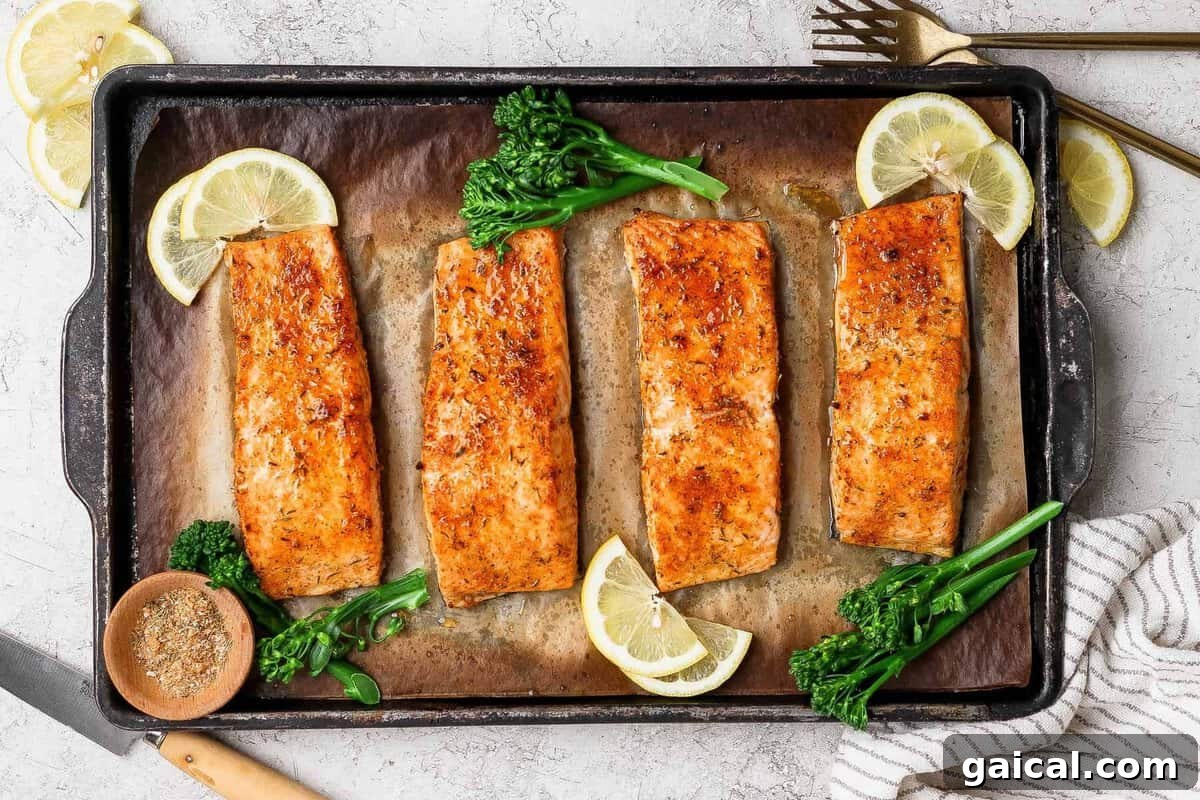 Broiled salmon fillets on a baking sheet garnished with lemon slices, broccolini, and a bowl of seasoning.