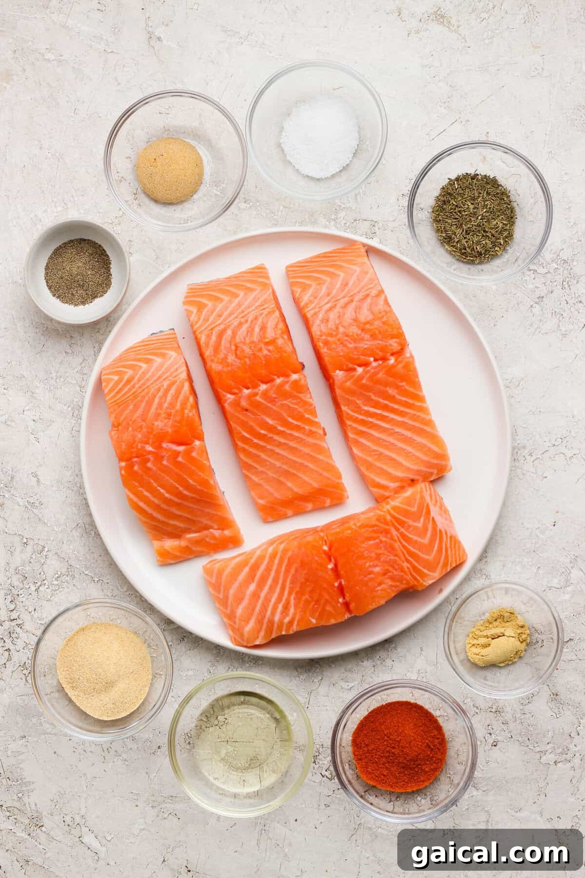 Raw salmon fillets on a plate surrounded by small bowls of seasoning, avocado oil, and herbs on a light countertop.