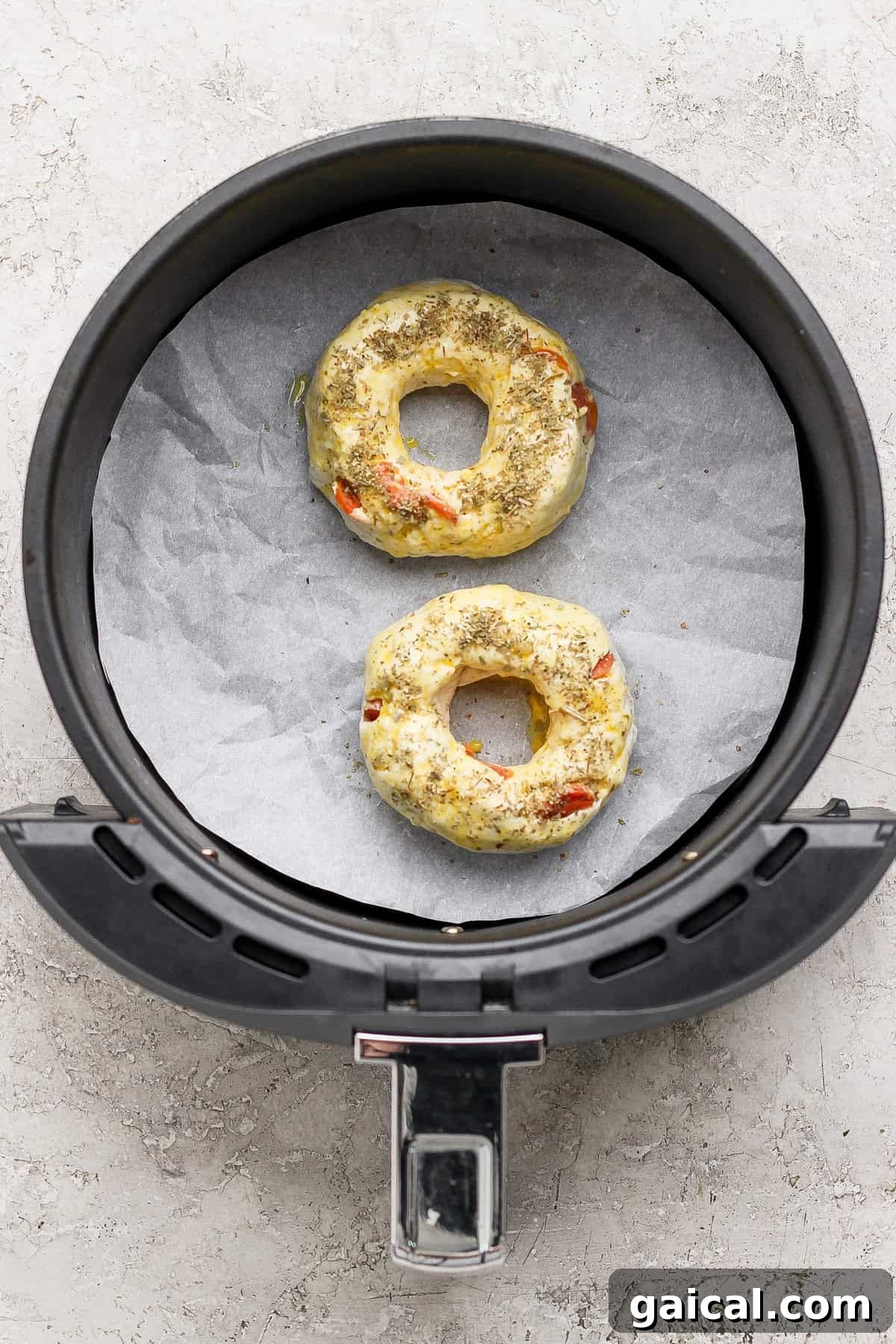Two uncooked pizza protein bagels with seasoning in an air fryer basket