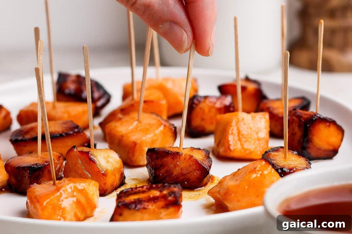 A hand picks up a toothpick with a piece of roasted sweet potato from a plate filled with several other sweet potato bites on toothpicks.