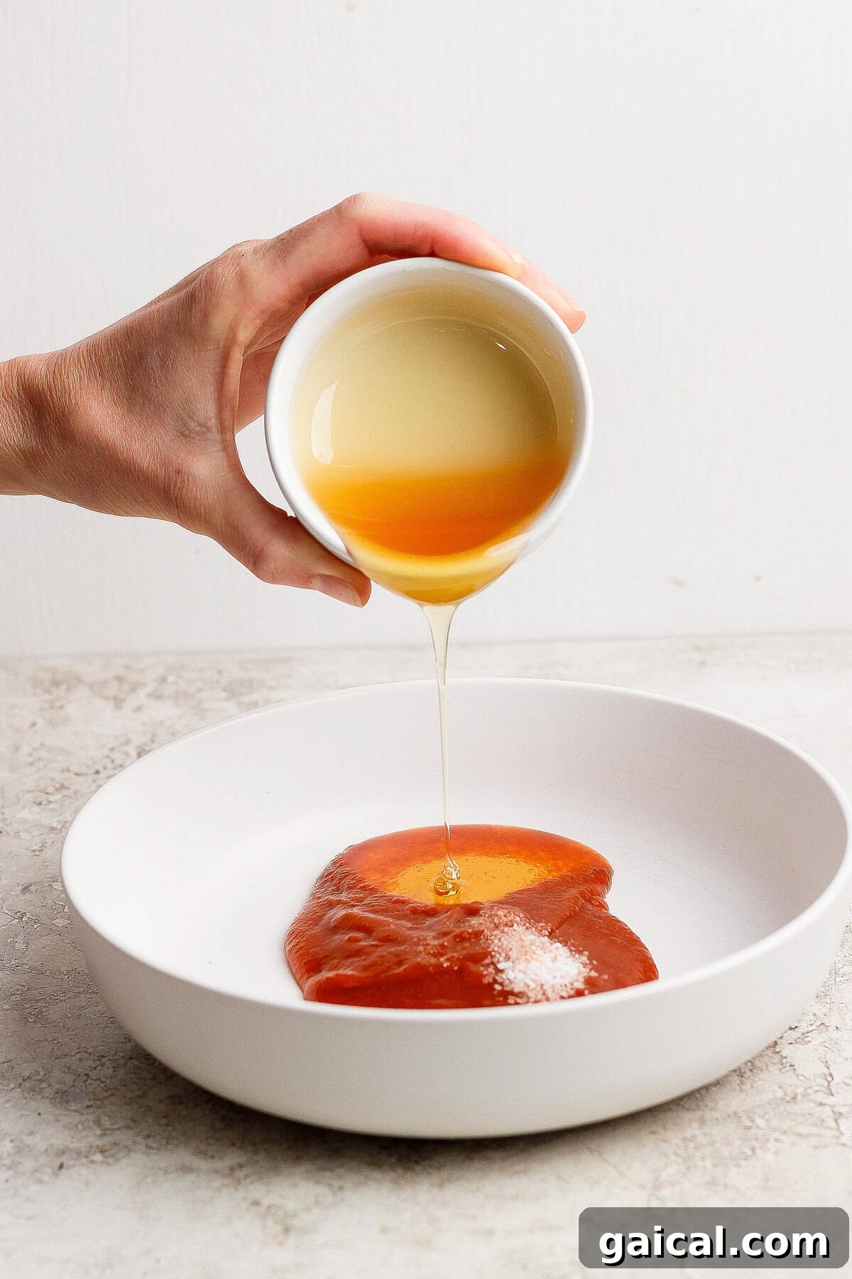 A hand pours honey from a small white bowl onto a plate of barbecue sauce.