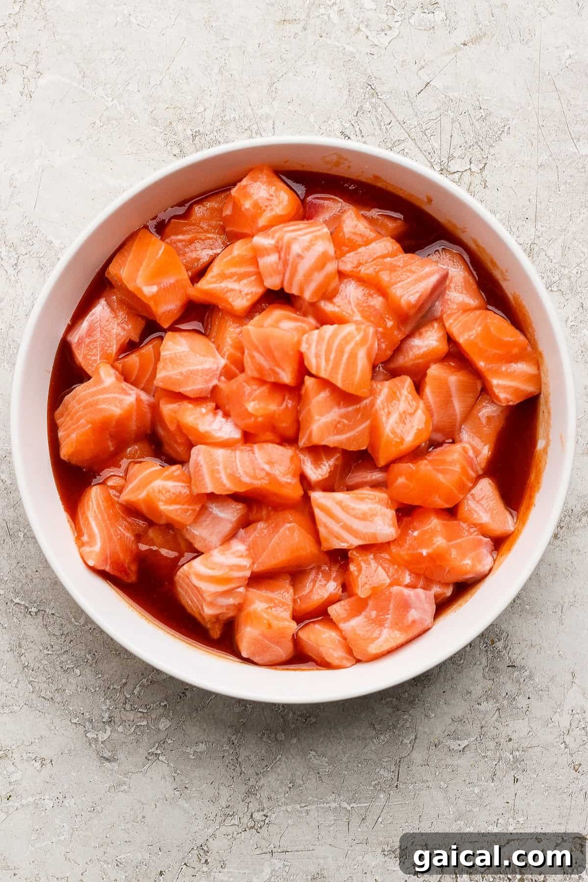 A white bowl filled with cubed raw salmon mixed with a red marinade, placed on a light textured surface.