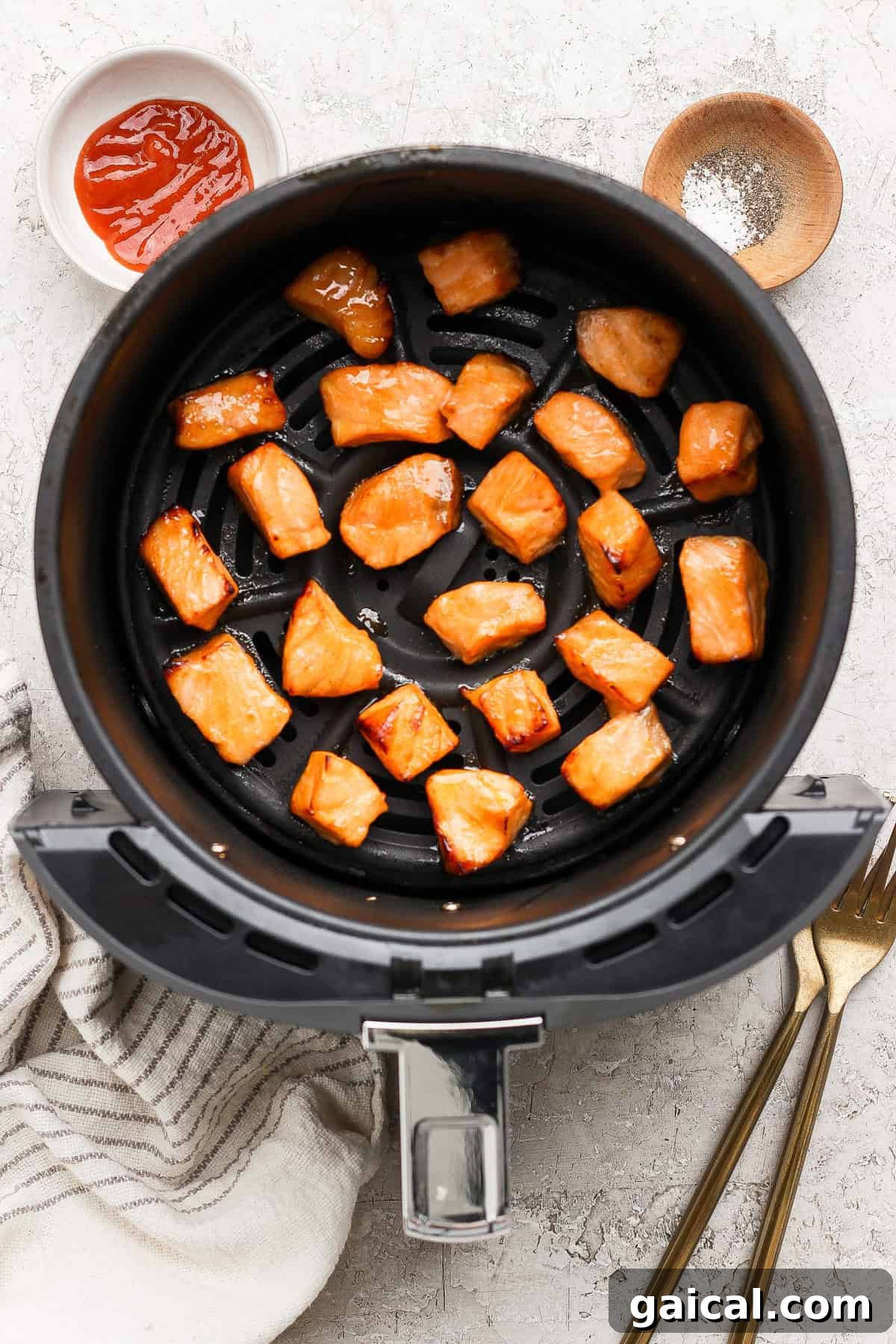 Cooked salmon pieces in an air fryer basket with bowls of sauce and seasoning nearby, a striped towel, and two forks on a light surface.