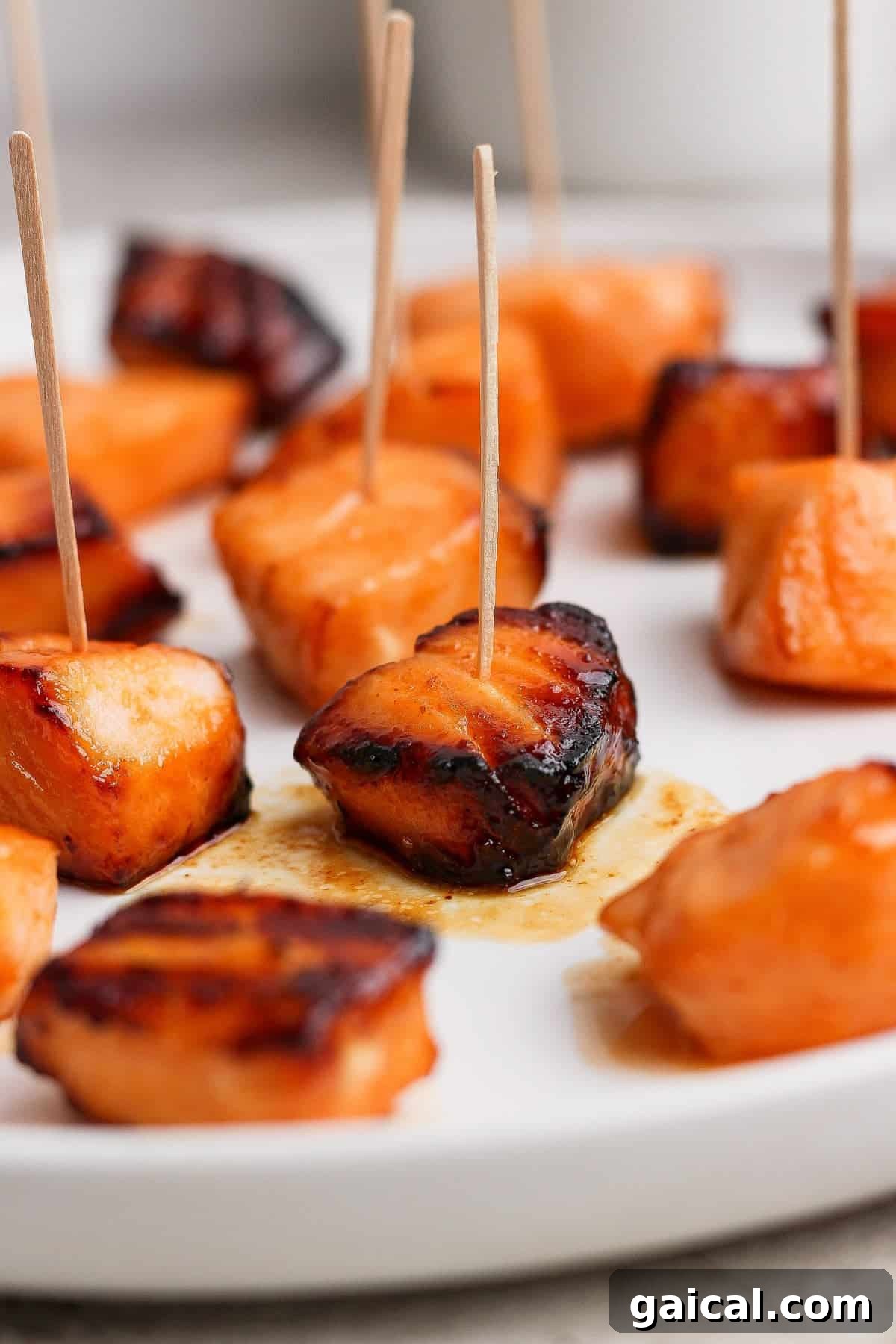Cubed pieces of glazed salmon, some with char marks, arranged on a white plate and skewered with wooden toothpicks.