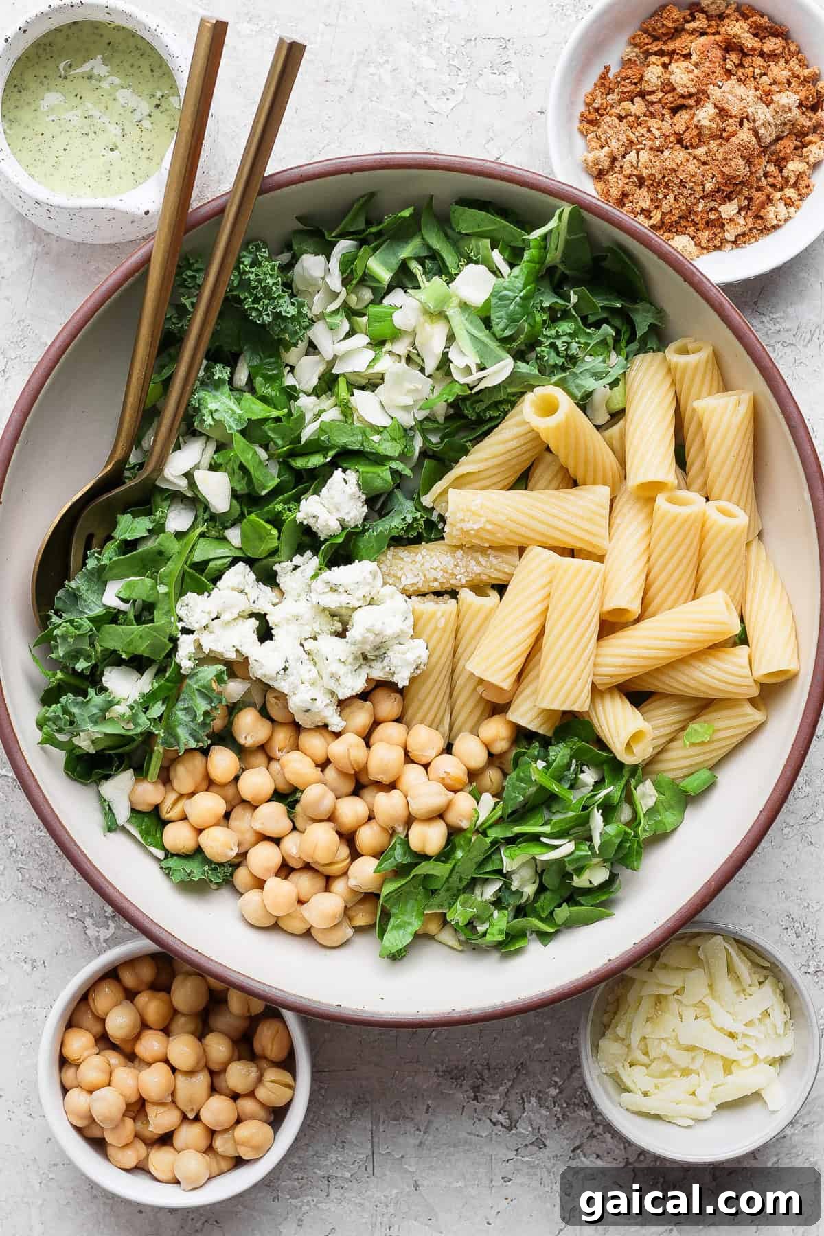 A large bowl with rigatoni pasta, chickpeas, leafy greens, feta cheese, and shaved parmesan, surrounded by bowls of dressing, crumbled topping, extra chickpeas, and parmesan.