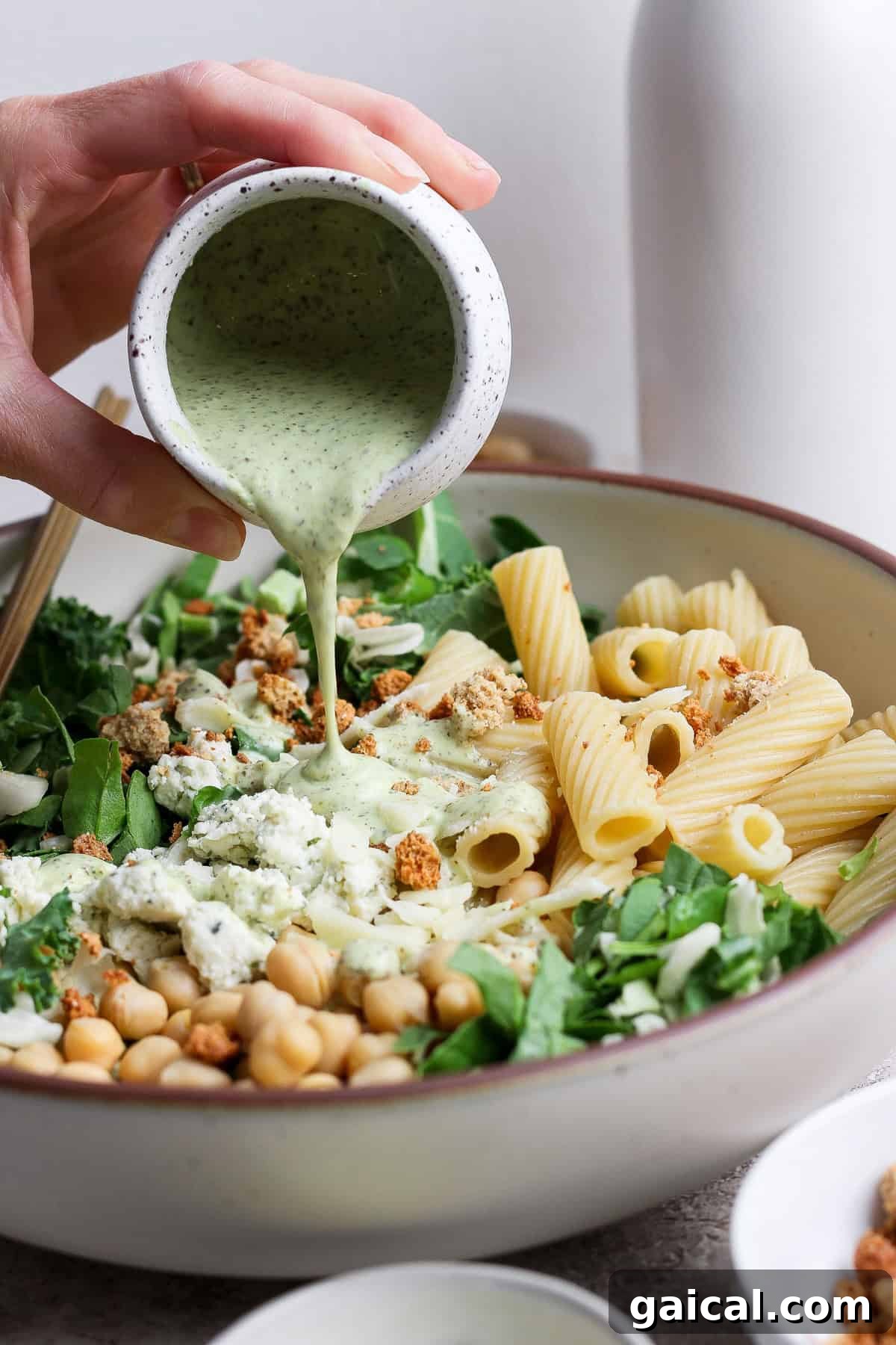 A hand pours creamy green dressing over a bowl of pasta salad with rigatoni, chickpeas, leafy greens, and crumbled toppings.