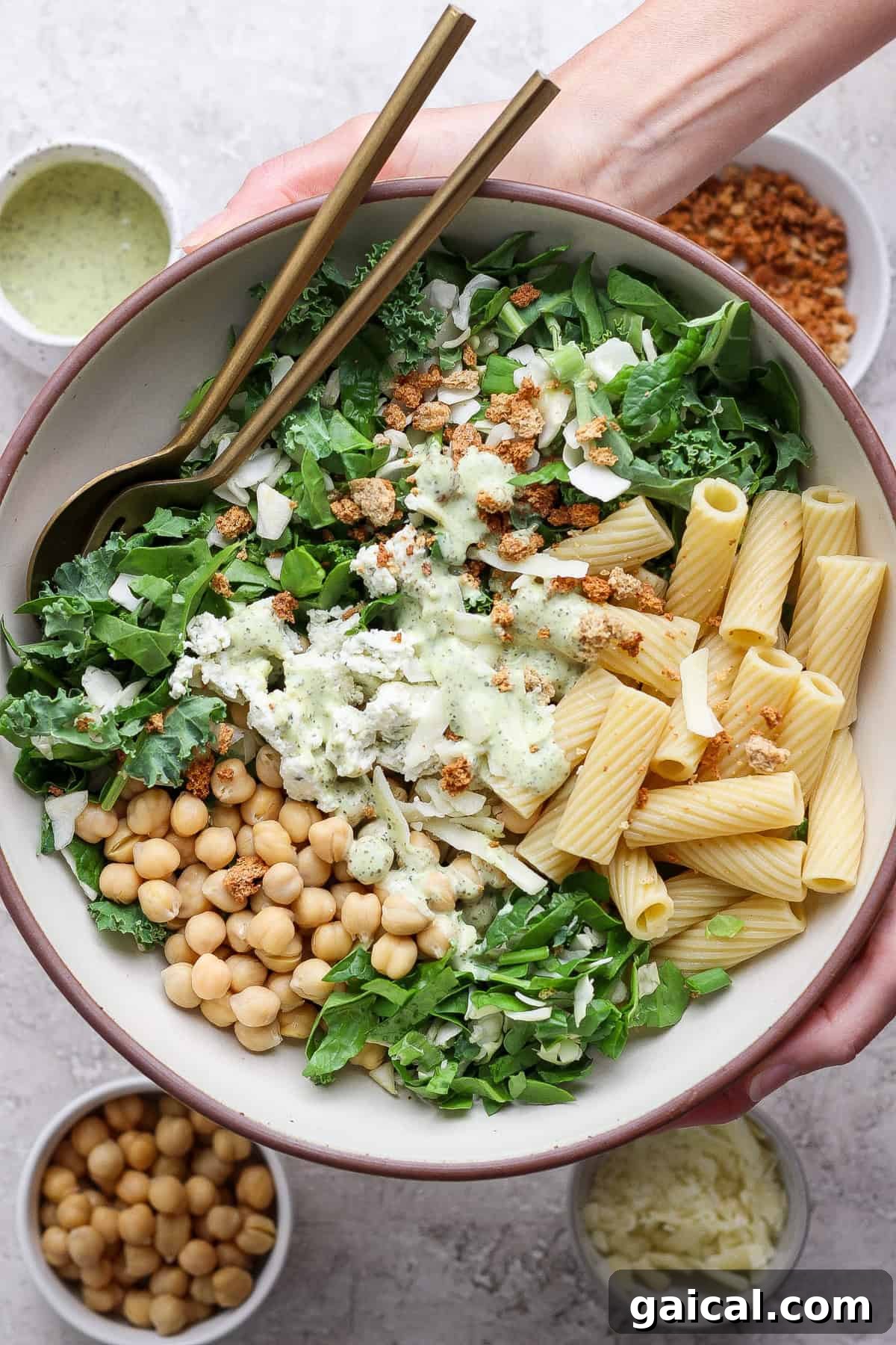 A bowl containing mixed greens, rigatoni pasta, chickpeas, cheese, and a creamy dressing, with two forks. Surrounding bowls hold extra chickpeas, shredded cheese, dressing, and crunchy toppings.