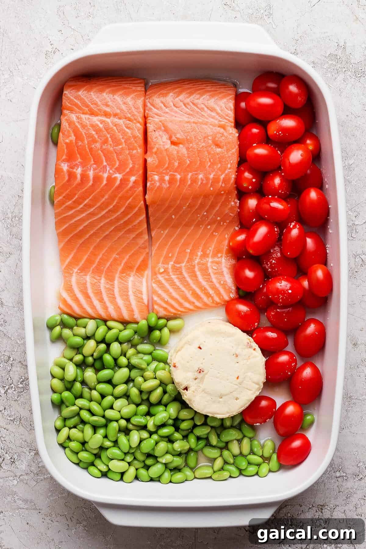 Raw salmon fillets, cherry tomatoes, edamame, and Boursin cheese assembled in a white casserole dish before baking