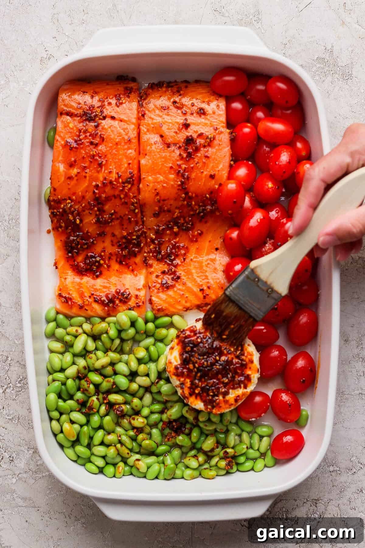 Brushing chili crunch onto Boursin cheese in a casserole dish with raw salmon and vegetables