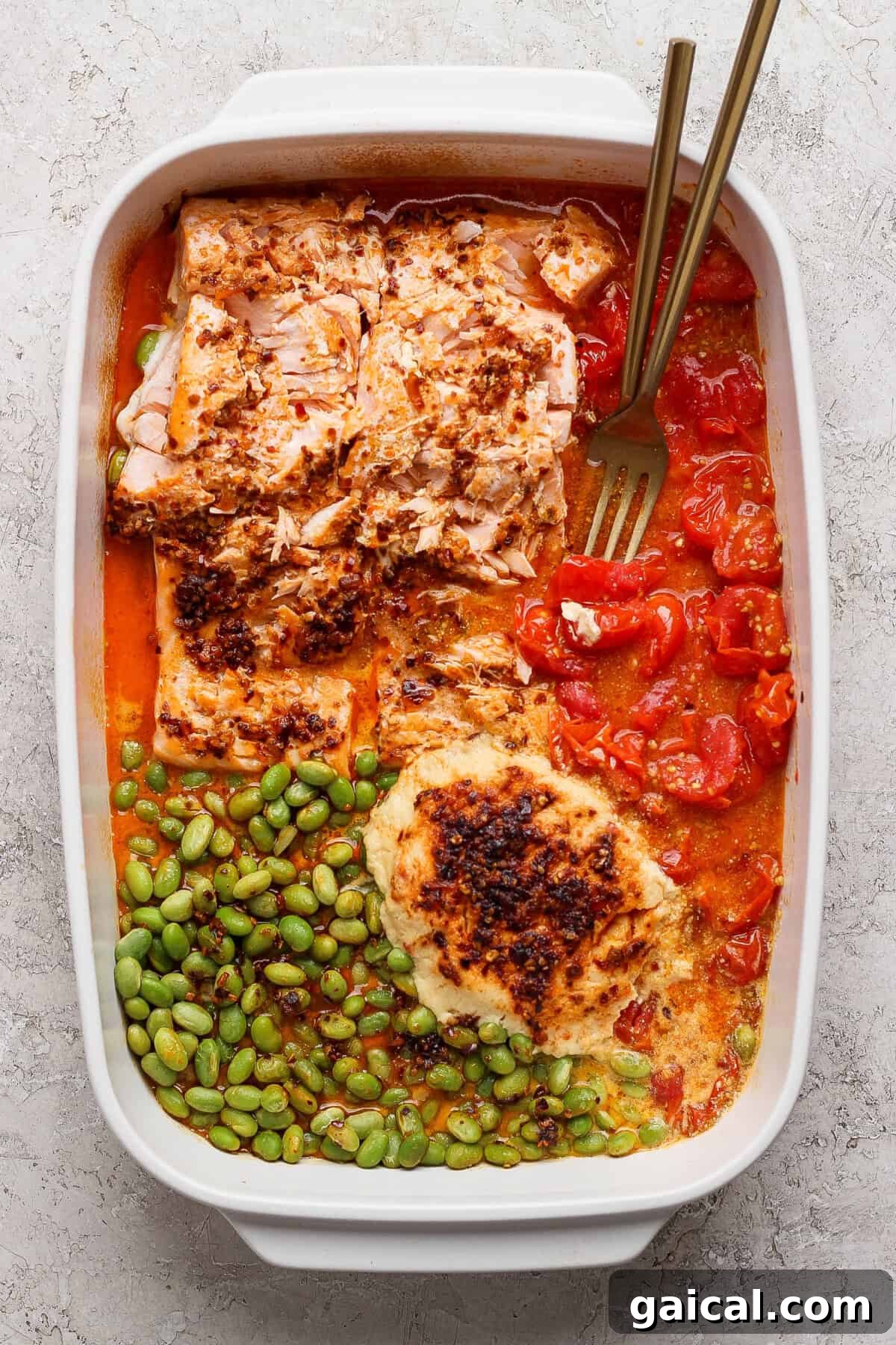 Baked salmon casserole with roasted tomatoes, edamame, and melted Boursin cheese with two gold forks in a white dish