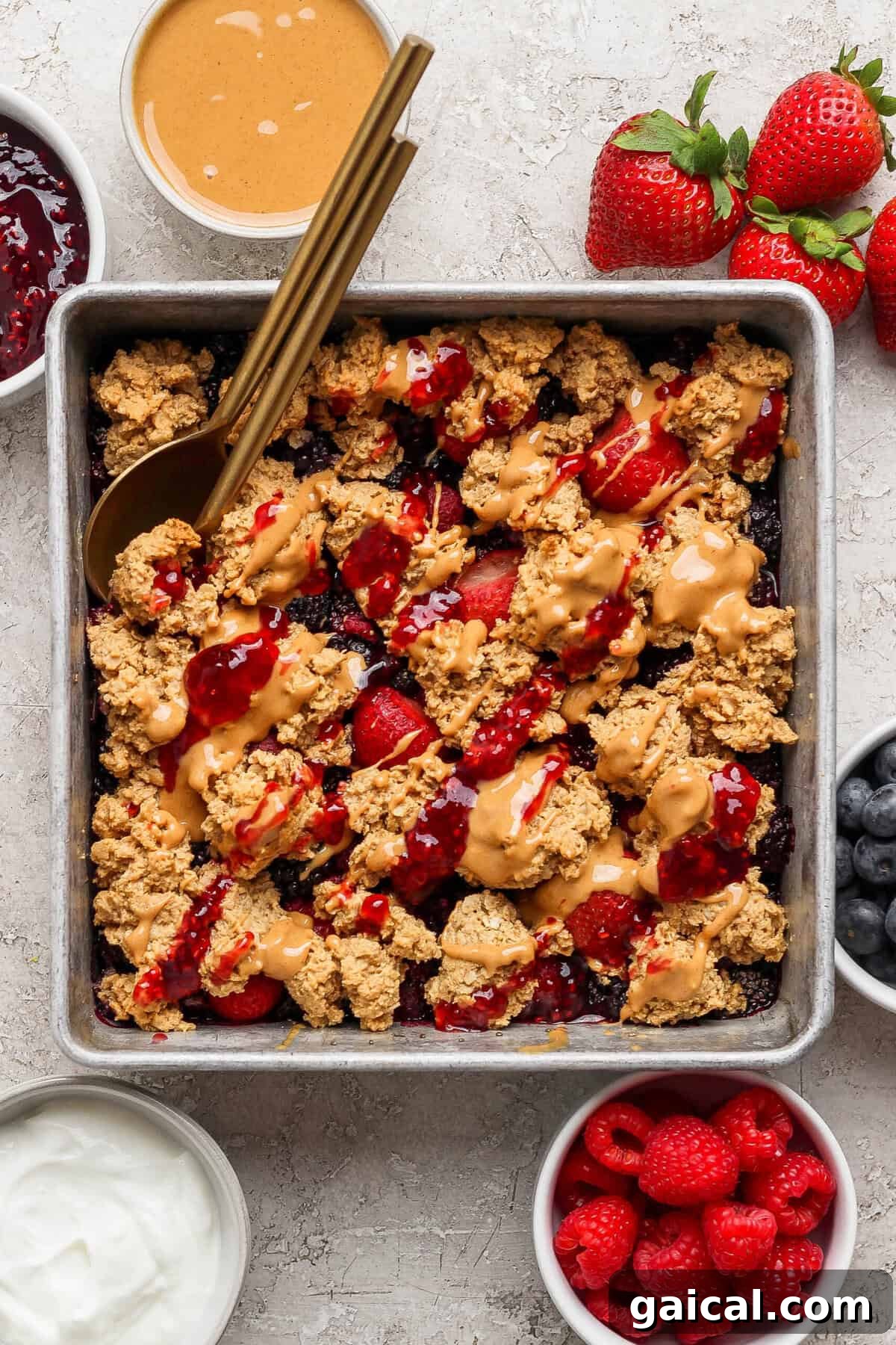 overhead view of protein berry crumble in a pan with peanut butter drizzle surrounded by bowls of jam yogurt and fresh berries