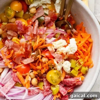 A salad bowl with shredded carrots, sliced red onions, cherry tomatoes, chickpeas, feta cheese, chopped bacon, and salami. Two forks are in the bowl.
