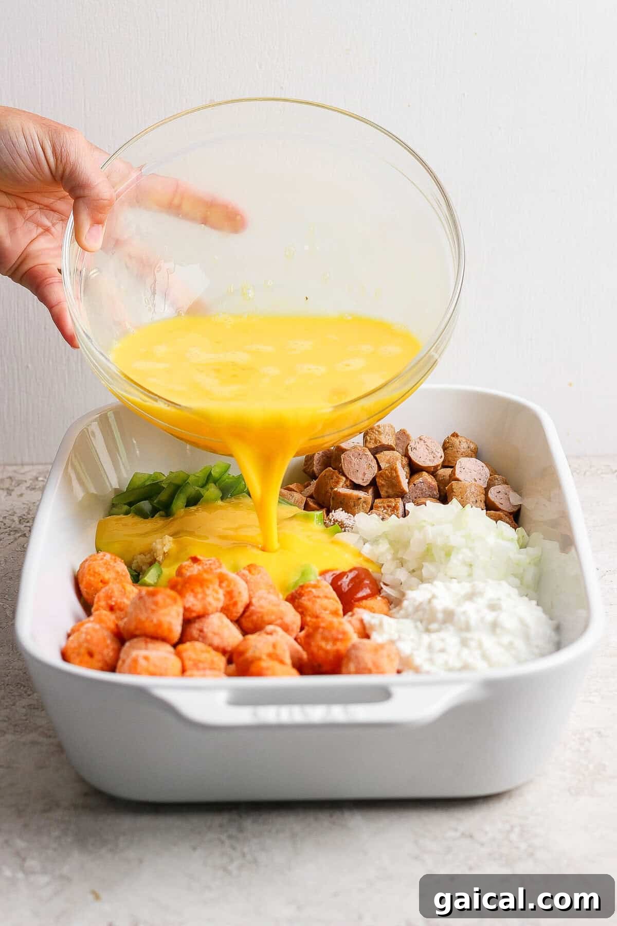 pouring whisked eggs over sweet potato tater tots and sausage in baking dish