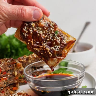 A hand dips a rectangular, sesame seed-topped pastry into a bowl of dark soy sauce.