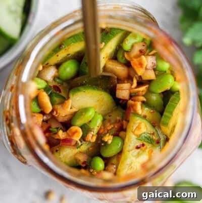 Top view of a jar filled with sliced cucumbers, edamame, chopped nuts, red onion, and spices, with a spoon inside.