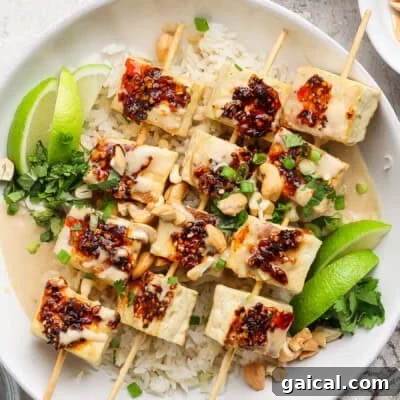 A dish with skewered tofu topped with chili sauce on rice, garnished with lime wedges, green onions, peanuts, and herbs.
