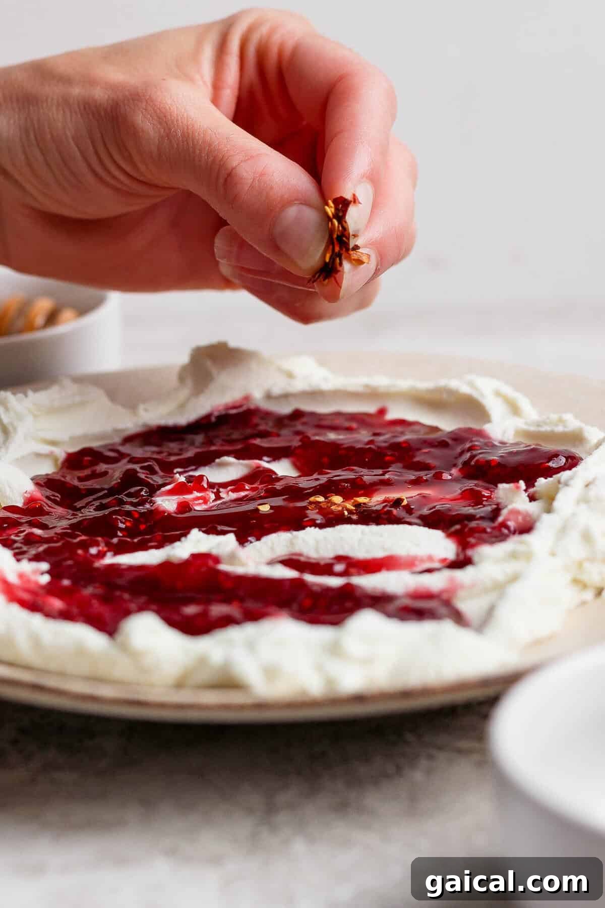 hand sprinkling red pepper flakes over goat cheese dip with raspberry jam