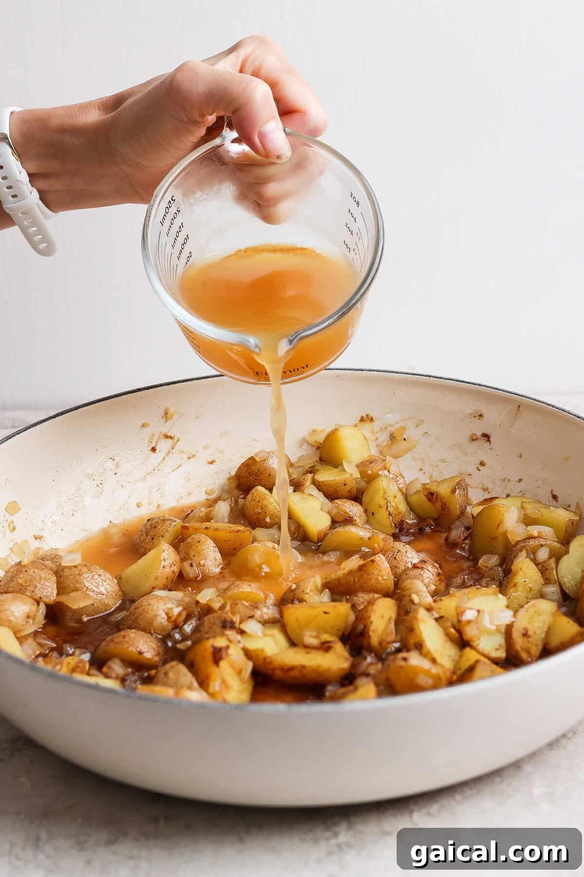 Apple cider being poured into a skillet of browned baby potatoes, onion, and garlic.