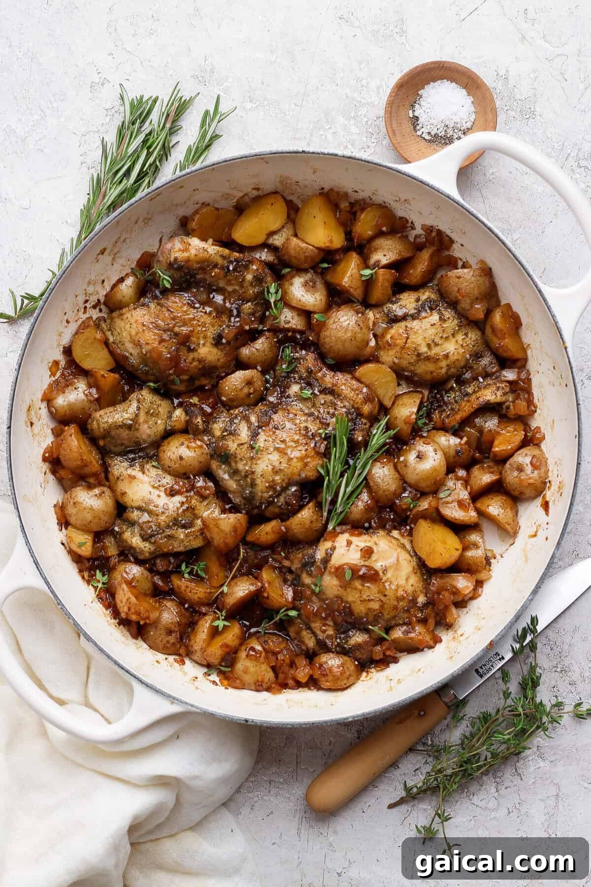 Overhead view of apple cider chicken thighs and baby potatoes in a white skillet with rosemary and thyme.