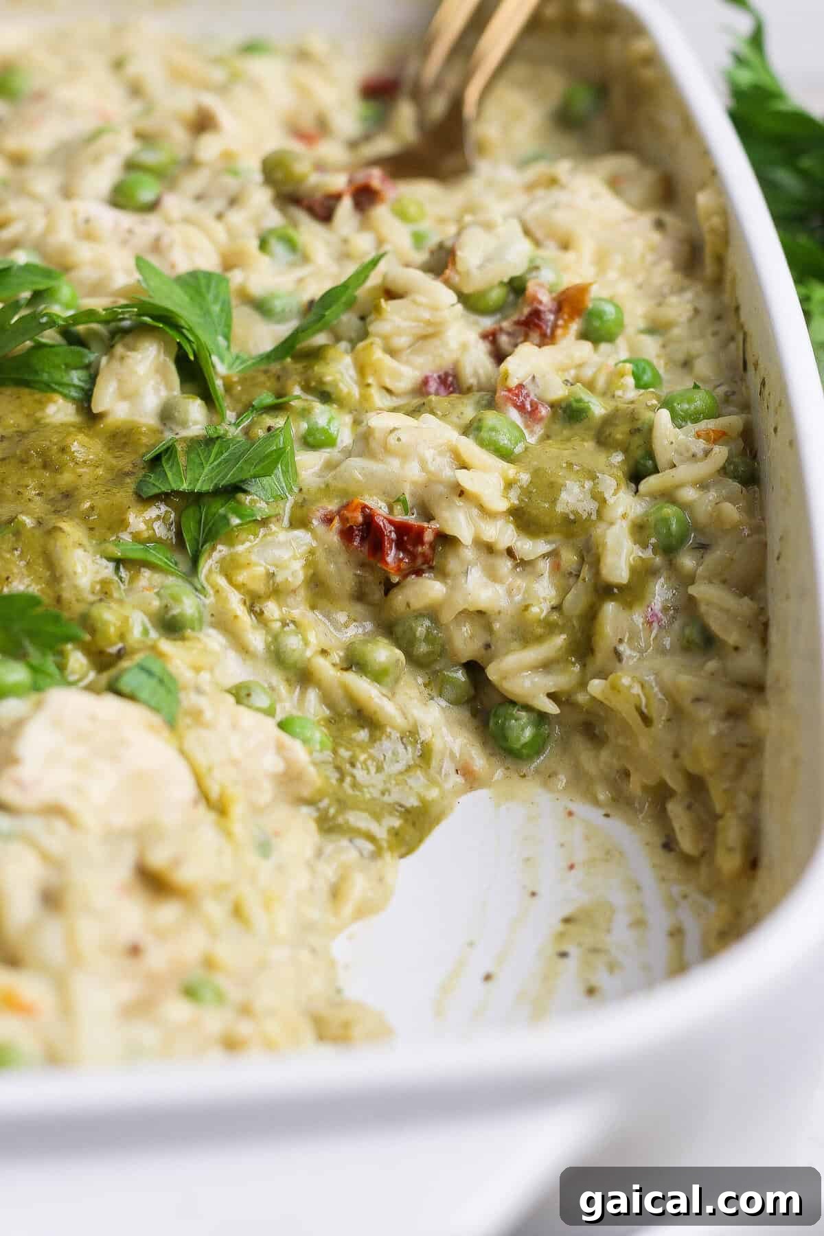 A serving spoon scooping creamy chicken pesto orzo with peas, sun-dried tomatoes, pesto, and fresh parsley from a white casserole dish.