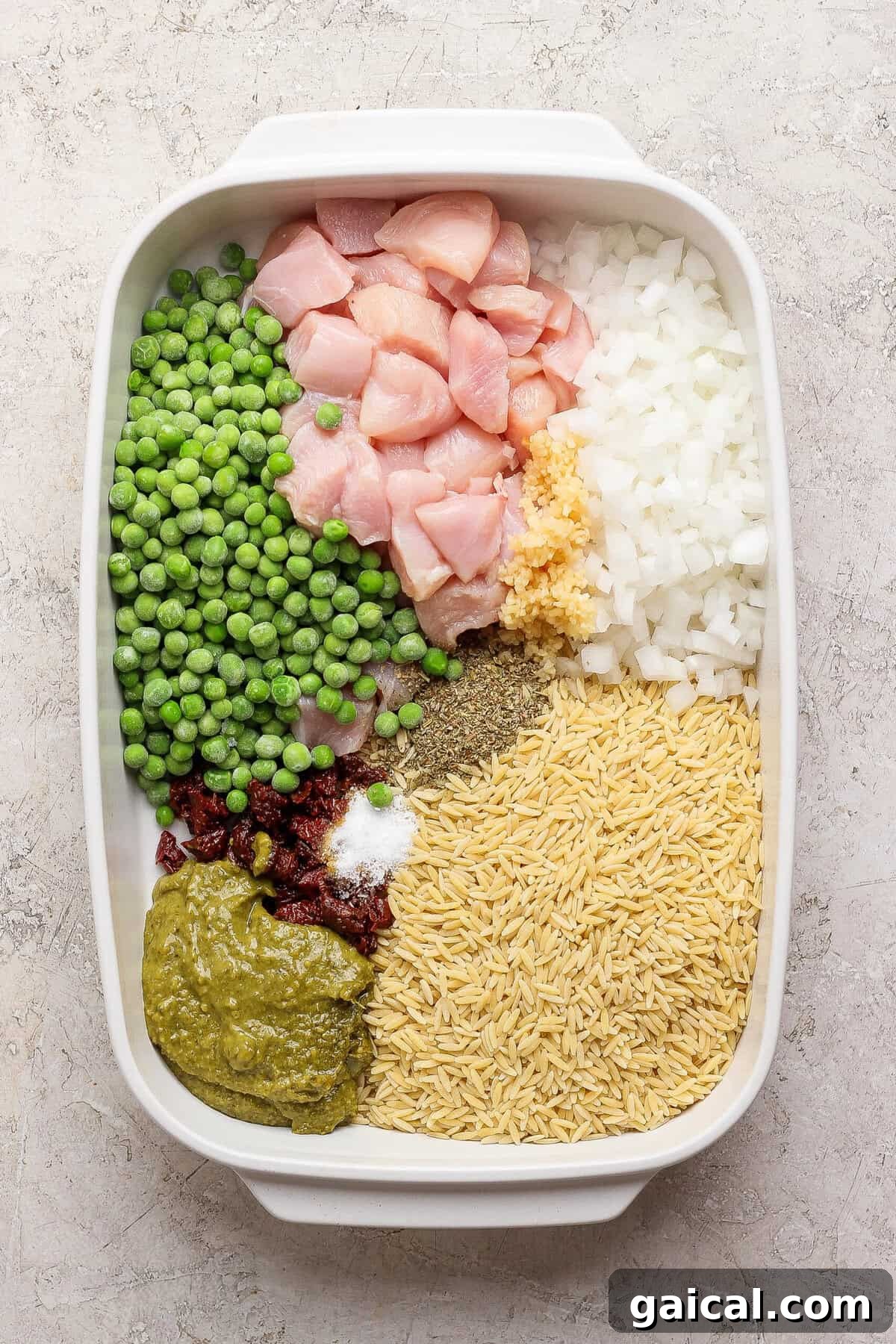 Raw chicken, dry orzo, frozen peas, onion, garlic, pesto, sun-dried tomatoes, and seasonings arranged in a white casserole dish.