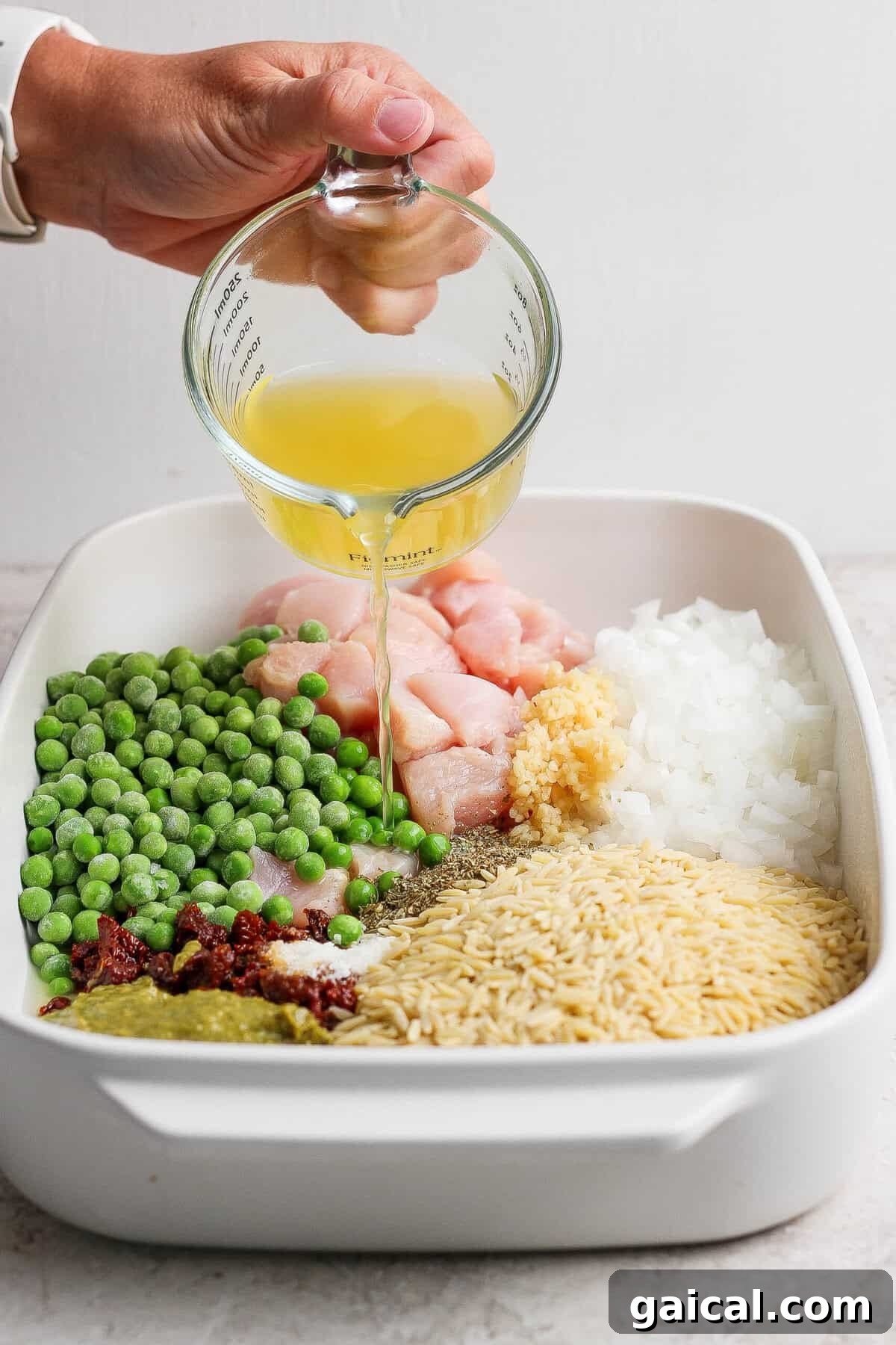 Chicken broth being poured over raw chicken, dry orzo, peas, pesto, sun-dried tomatoes, onion, garlic, and seasonings in a casserole dish.