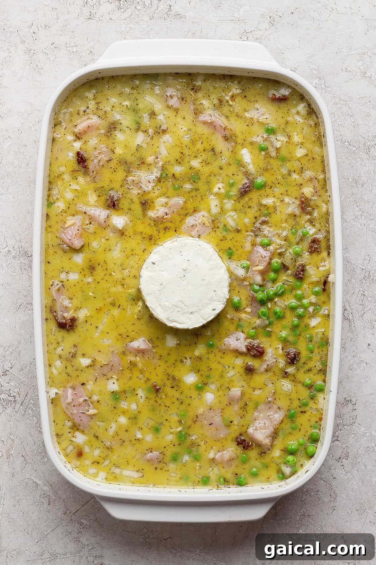 Unbaked chicken pesto orzo mixture in a white casserole dish with a whole puck of Boursin in the center.