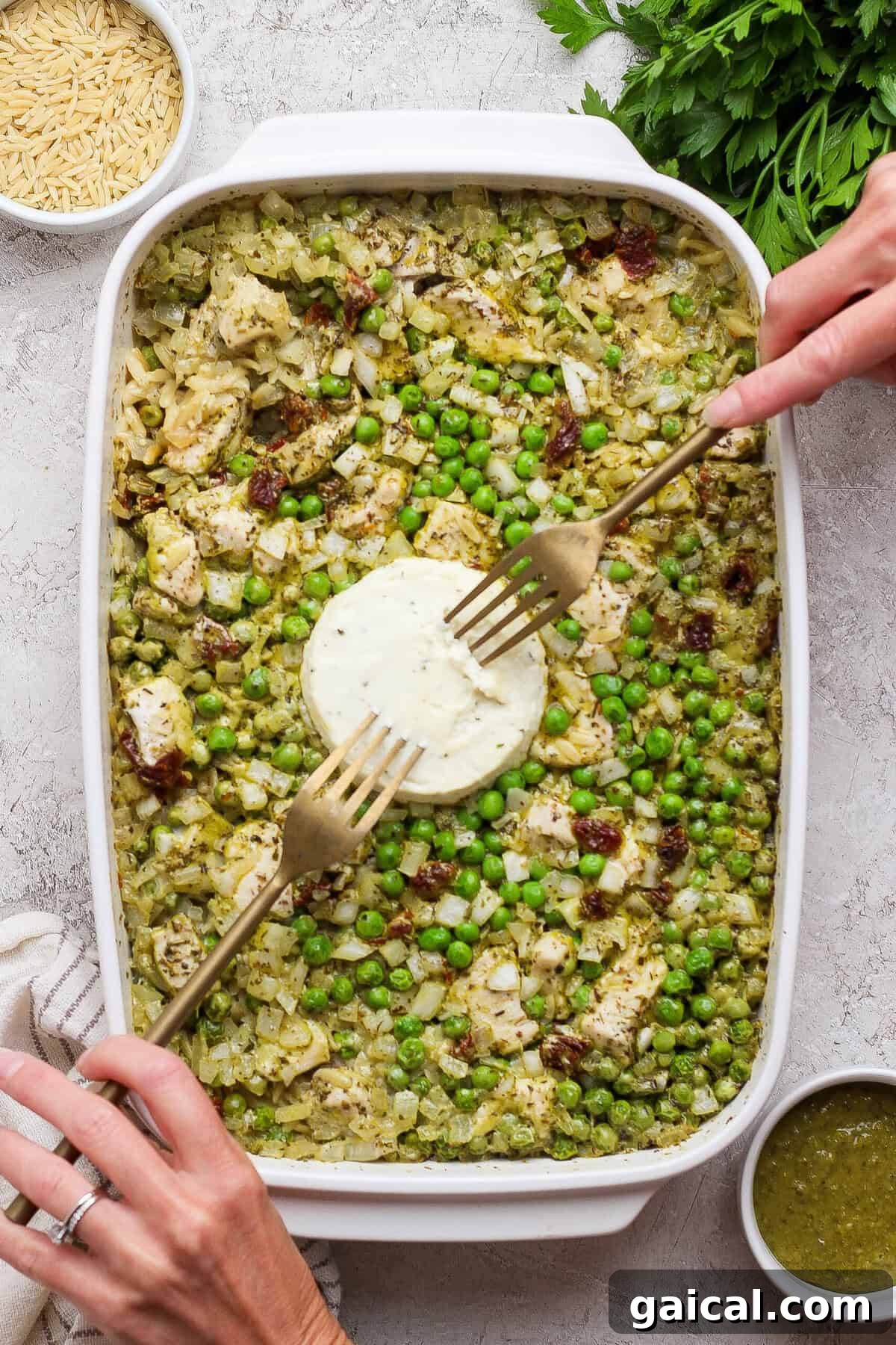 Hands using forks to mix softened Boursin into baked chicken pesto orzo with peas and sun-dried tomatoes.