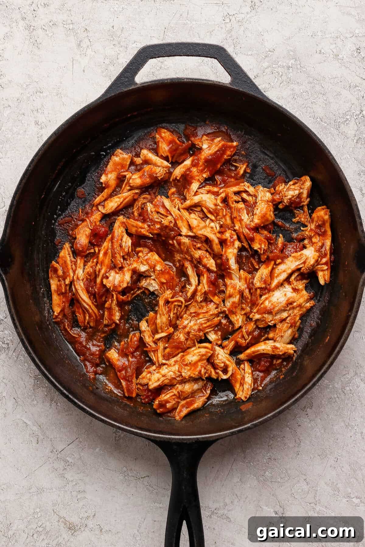 Shredded cooked chicken coated in red taco cooking sauce in a cast-iron skillet.