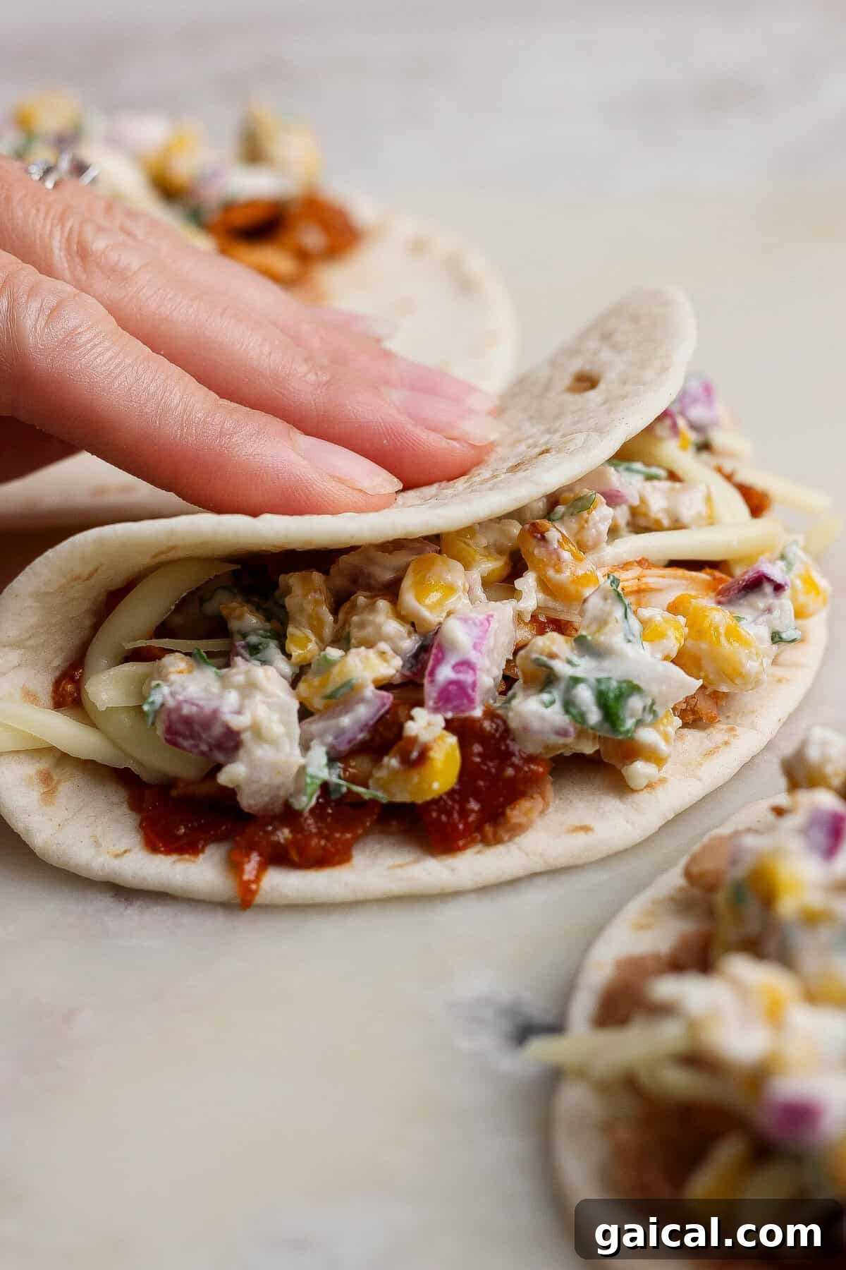 A hand folding a filled flour tortilla over street corn salad, shredded chicken, refried beans, and cheese.