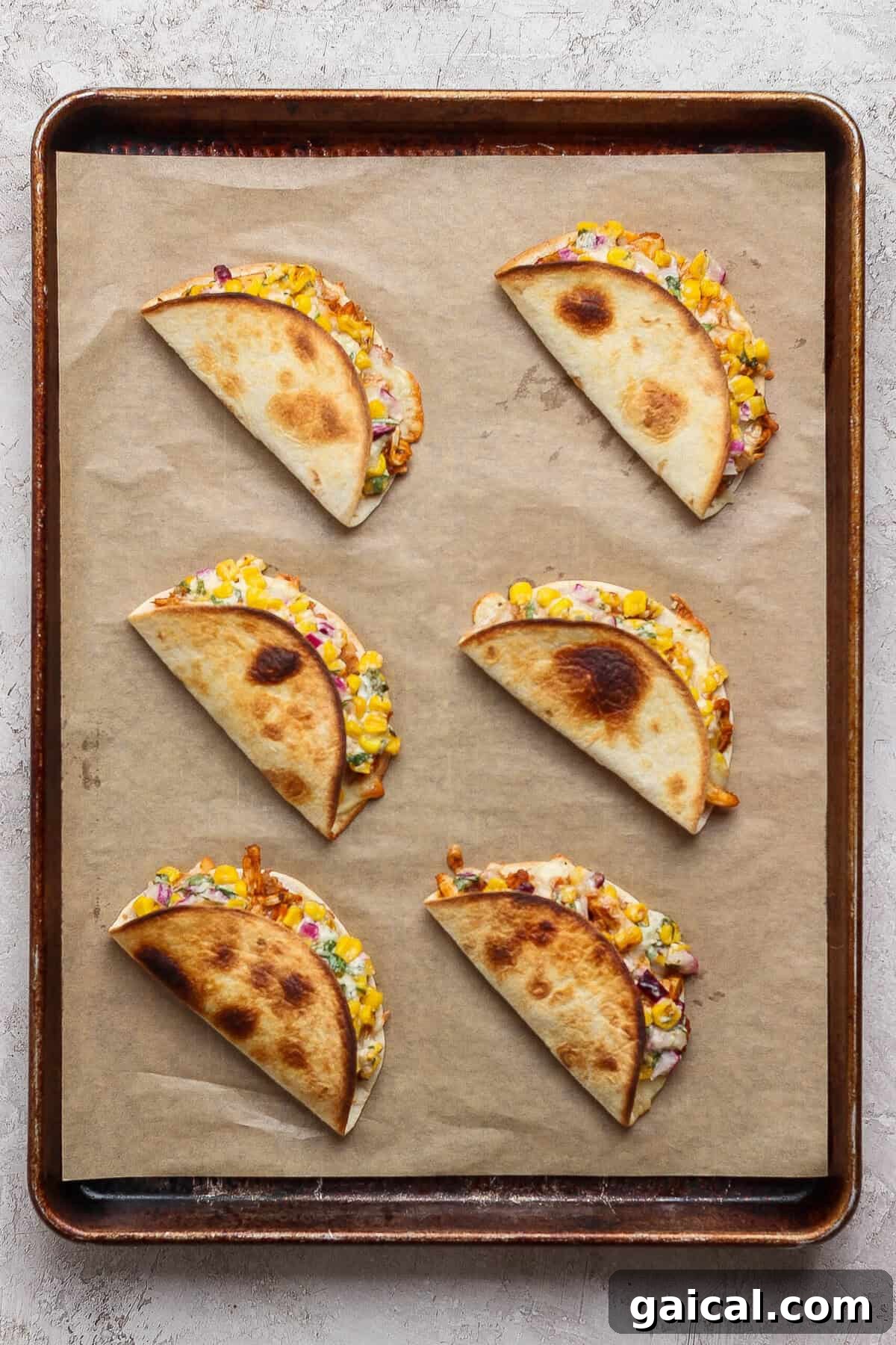 Six folded street corn chicken tacos baked until golden on a parchment-lined metal sheet pan.