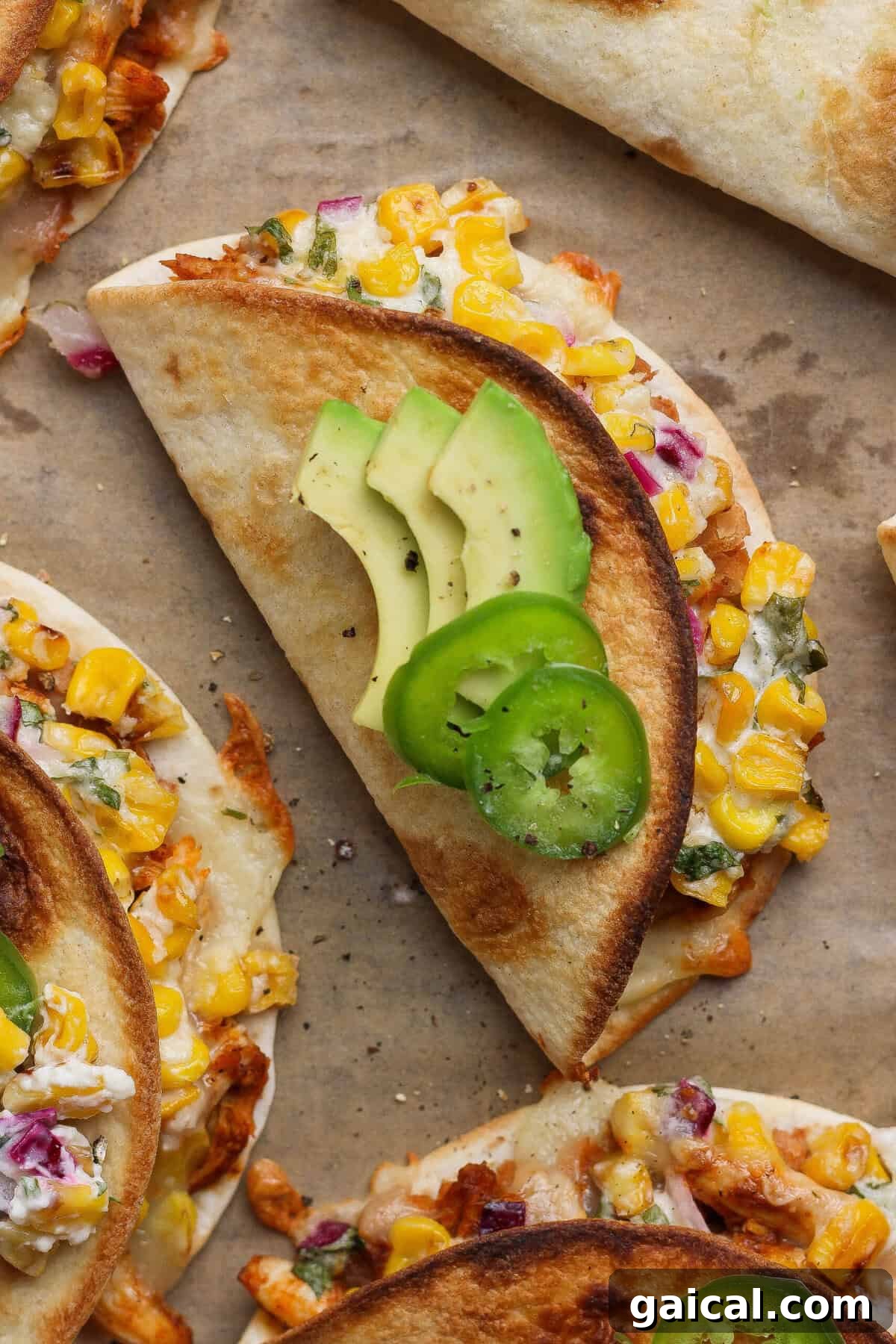 Crispy street corn chicken tacos topped with sliced avocado and jalapeño on a parchment-lined baking sheet.